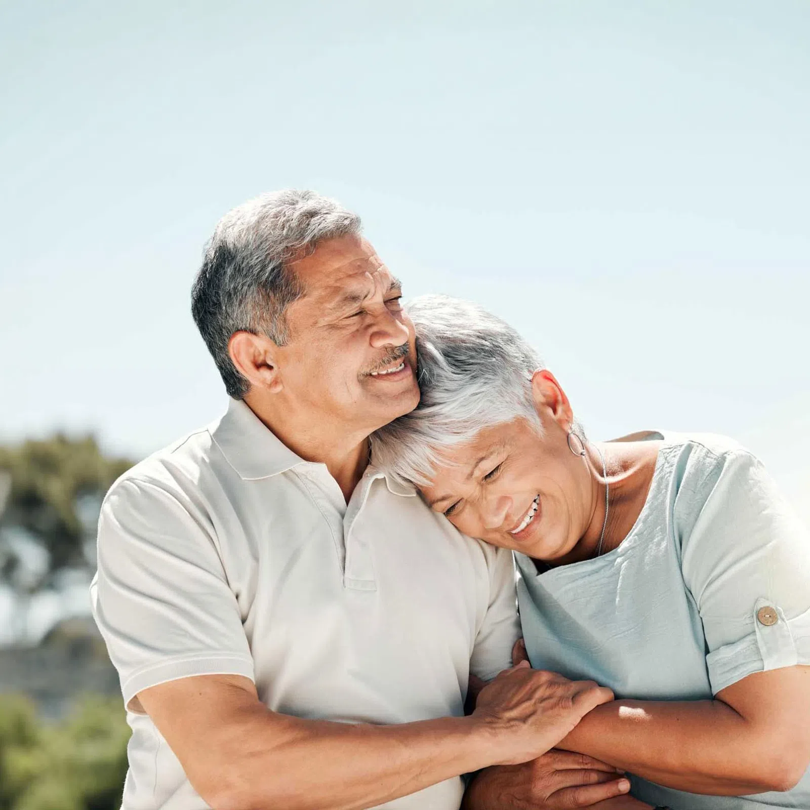 Smiling senior couple embracing outdoors with a bright, clear sky and blurred greenery background.