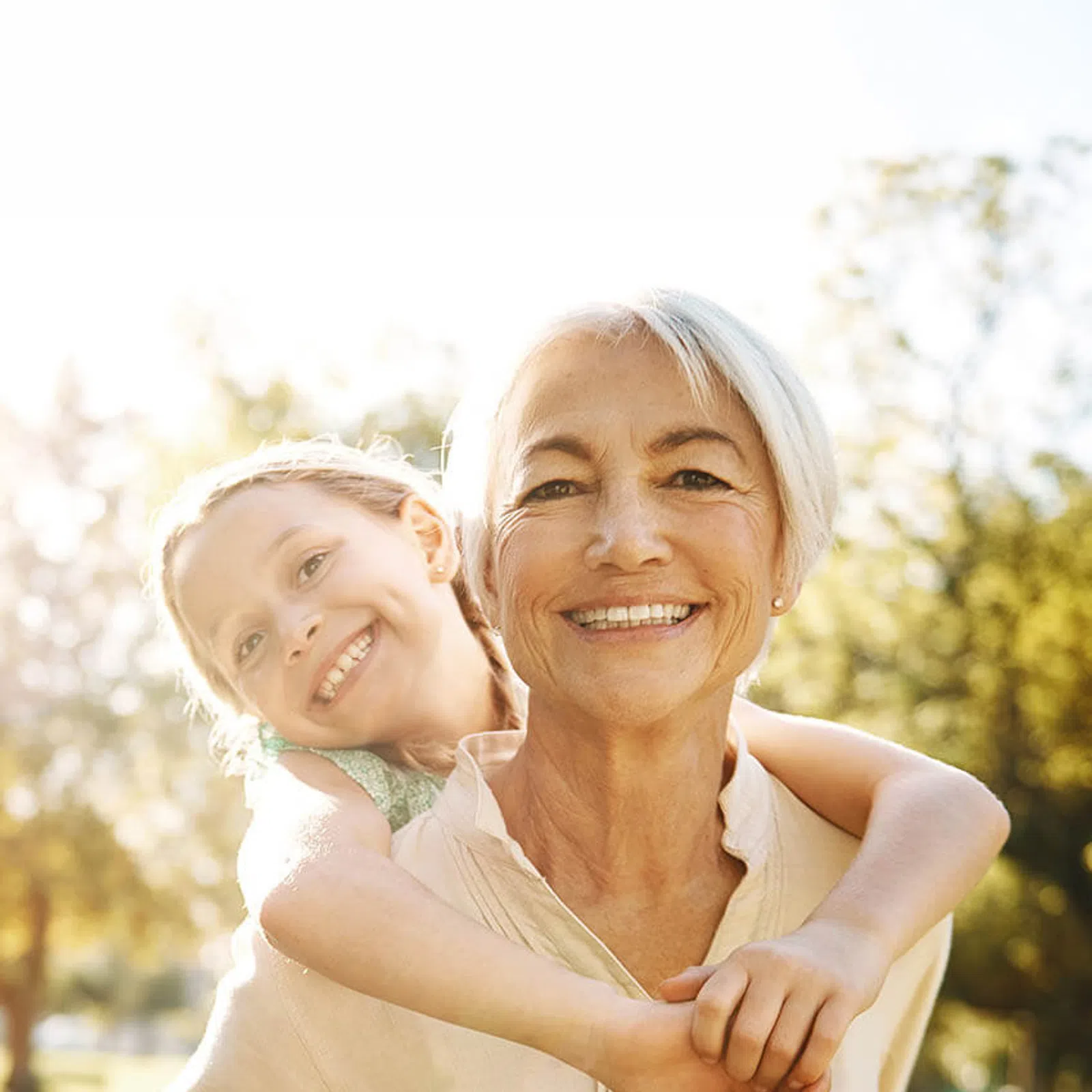 Smiling elderly woman with short white hair carrying a happy young girl on her back outdoors in a sunlit park