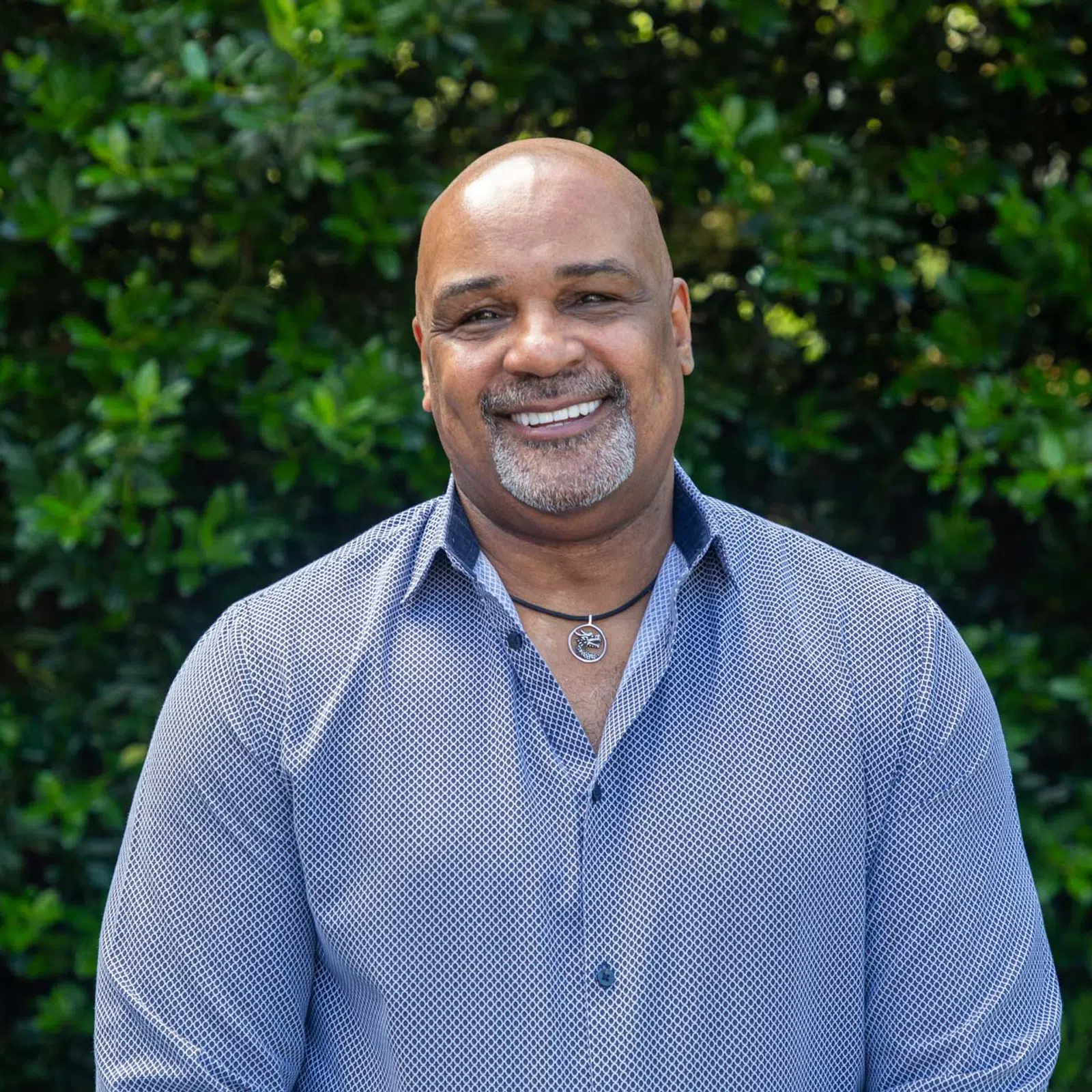 Smiling bald man wearing a blue patterned shirt and necklace stands outdoors against green foliage background