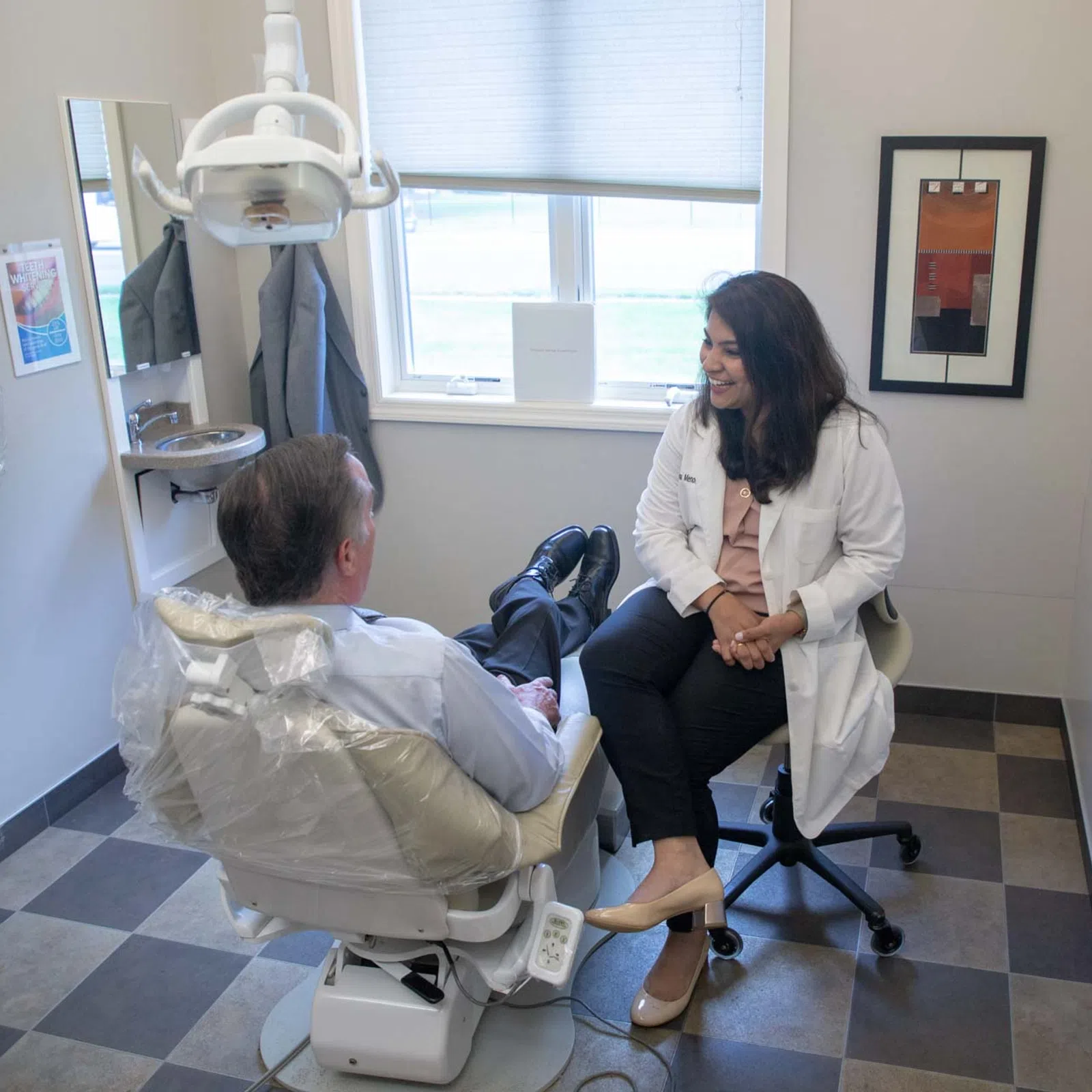 Dentist in white coat consults with male patient sitting in dental chair in bright clinic room.