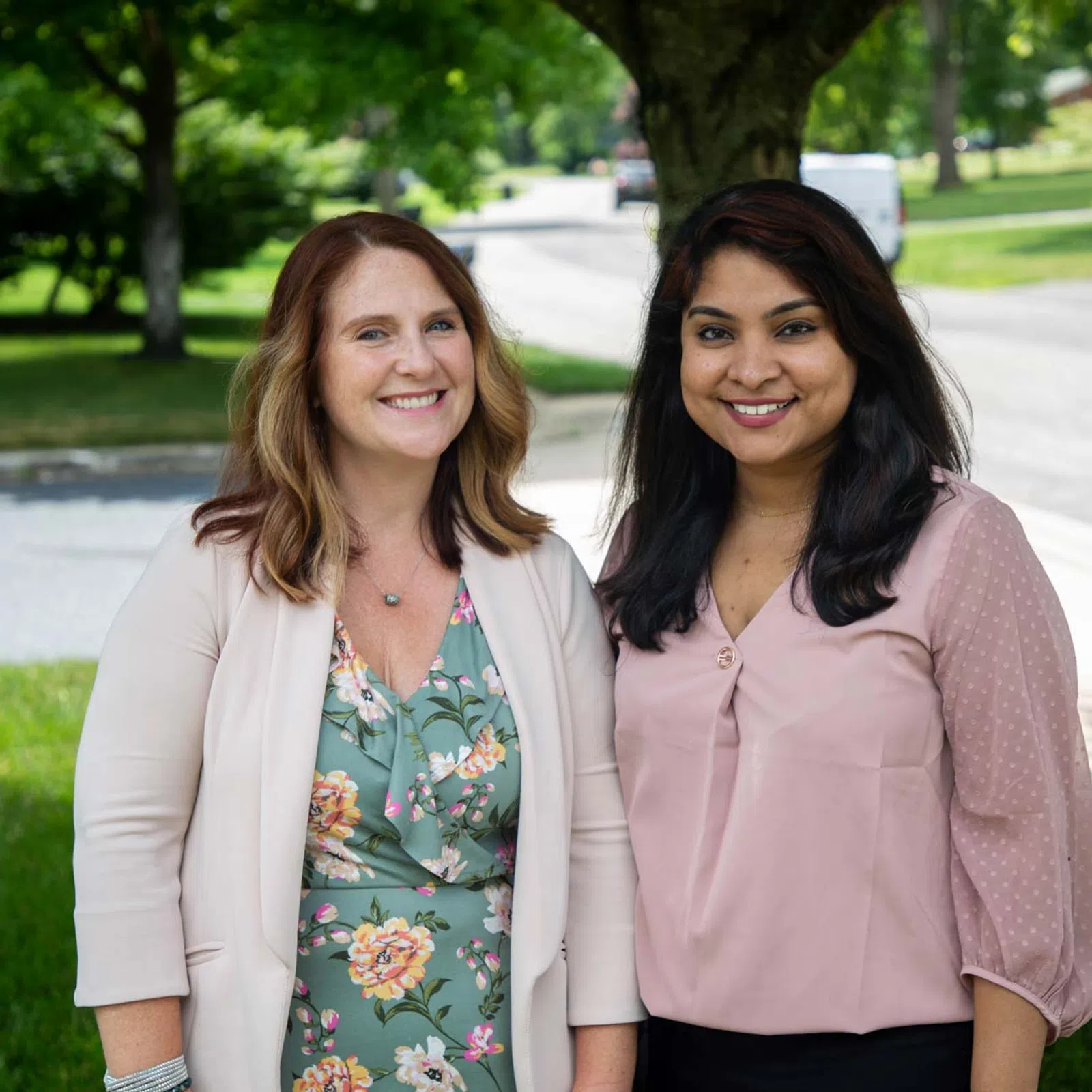Two smiling women standing outdoors under a tree on a sunny day with neighborhood street background