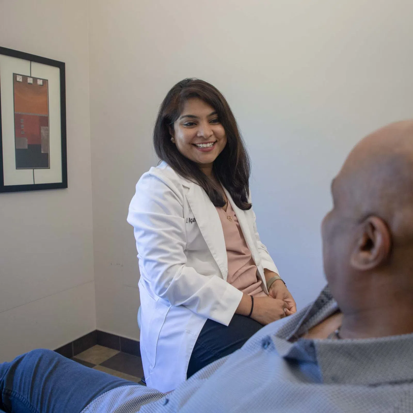 Female doctor in white coat smiling and consulting with male patient in medical office setting.