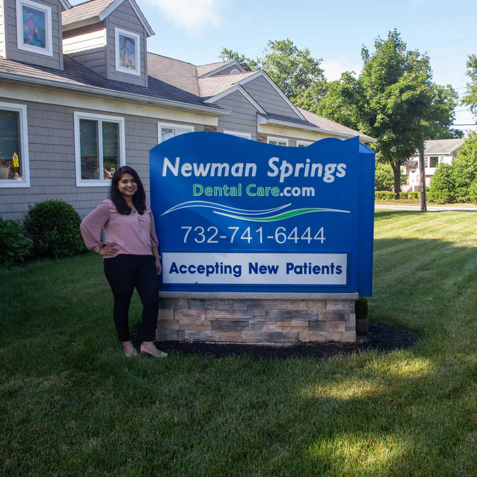 Woman standing next to Newman Springs Dental Care sign advertising new patients and phone number outdoors.