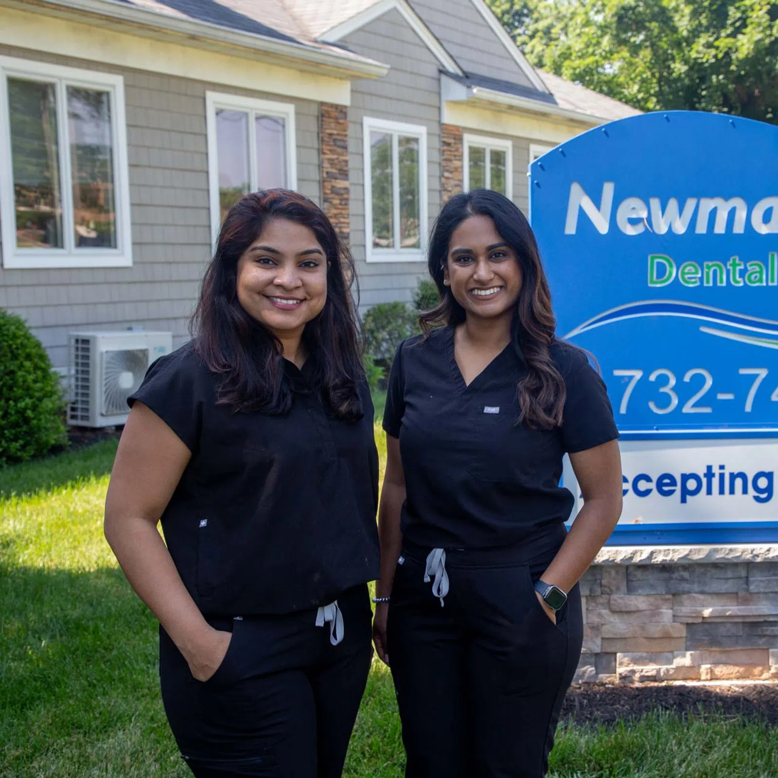 Two smiling women in black scrubs stand outside Newman Springs Dental Care with a sign offering new patients.