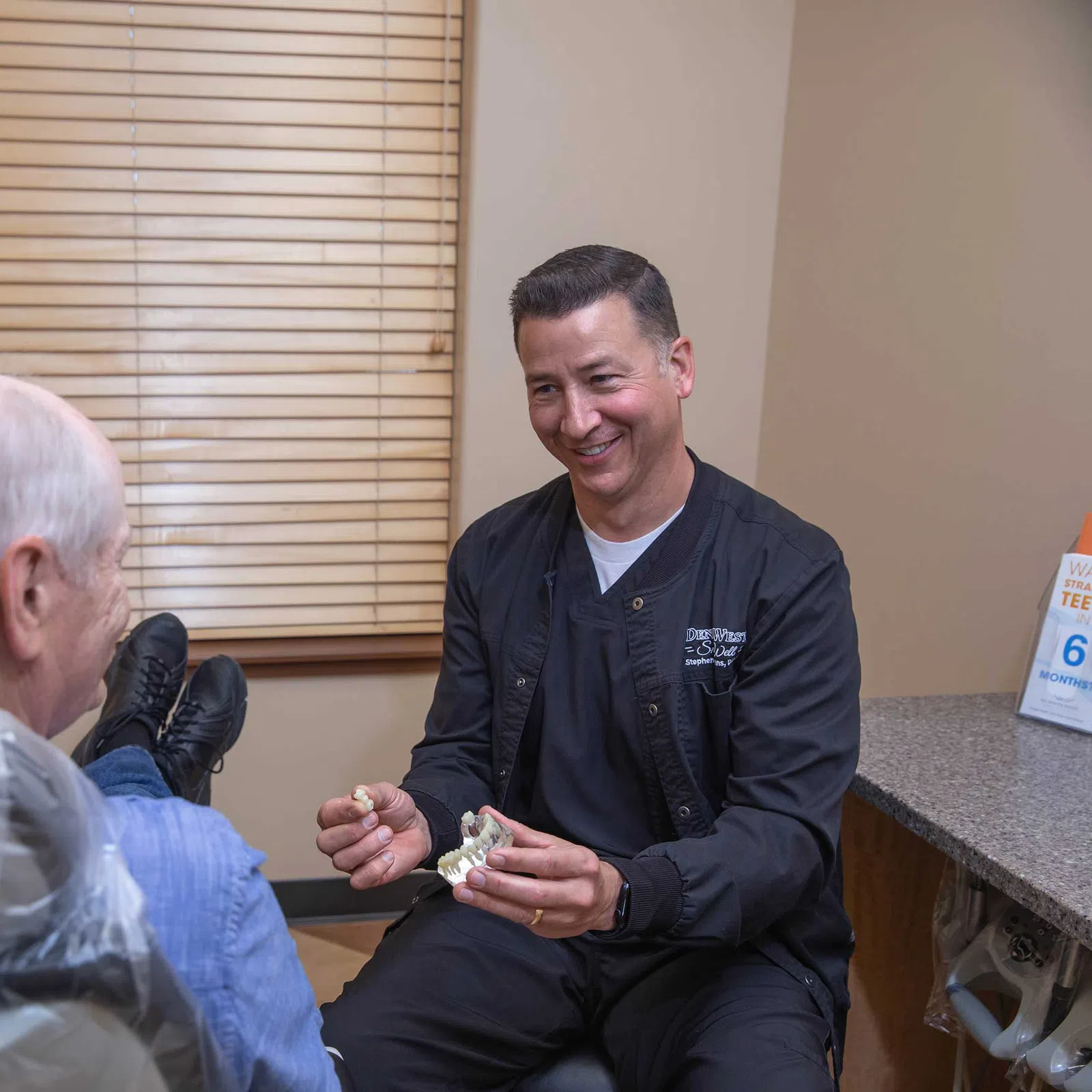 Dentist explaining dental model to elderly male patient in a clinic with beige walls and blinds.