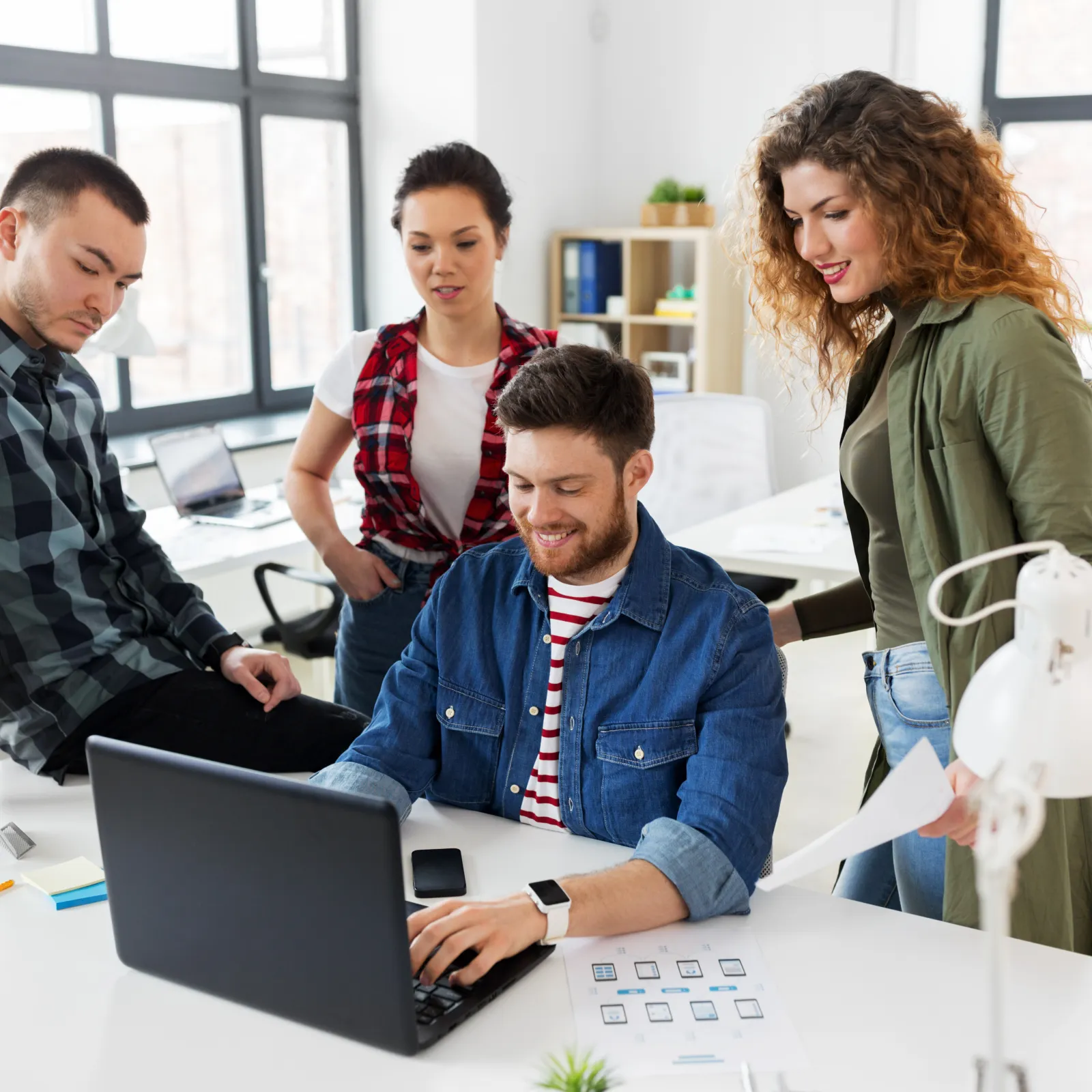Four young professionals collaborating around a laptop in a bright modern office with large windows.