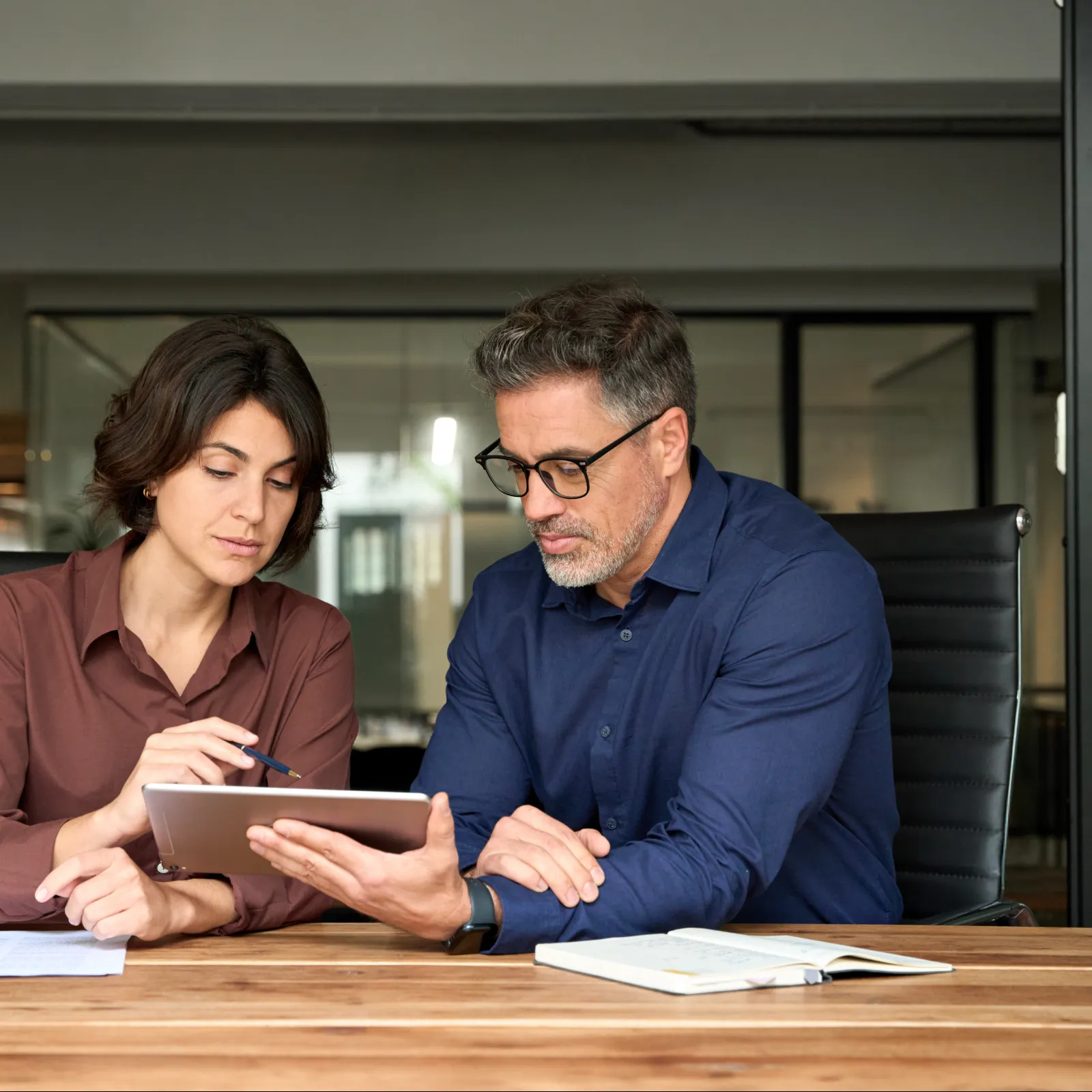 Business colleagues discussing data on a tablet in a modern office setting with documents on the table.