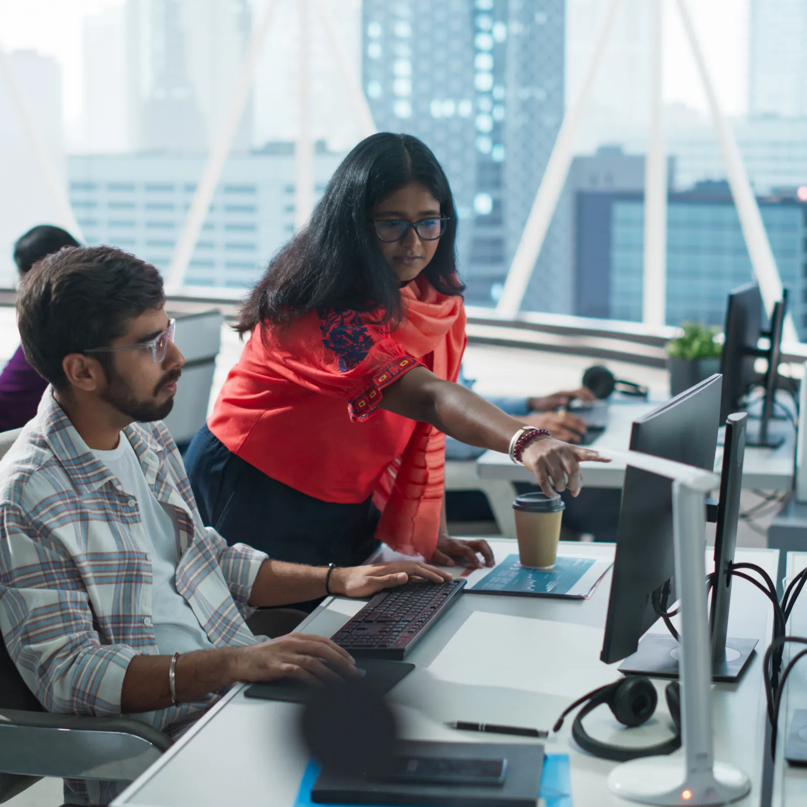 Two colleagues collaborate on a computer in a modern office with city views and multiple monitors.