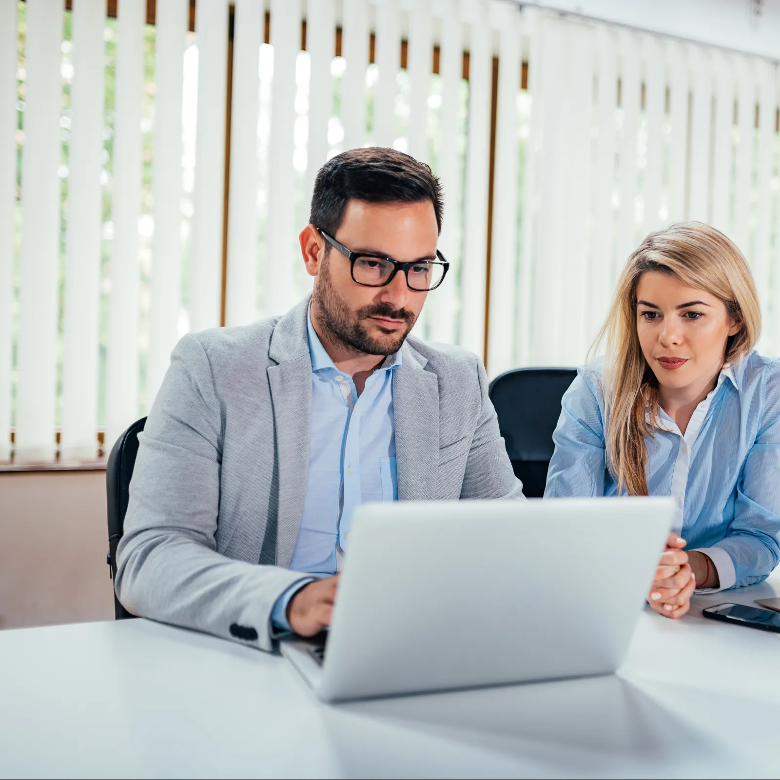 Two professionals in business attire working together on laptops at a bright office with vertical blinds.