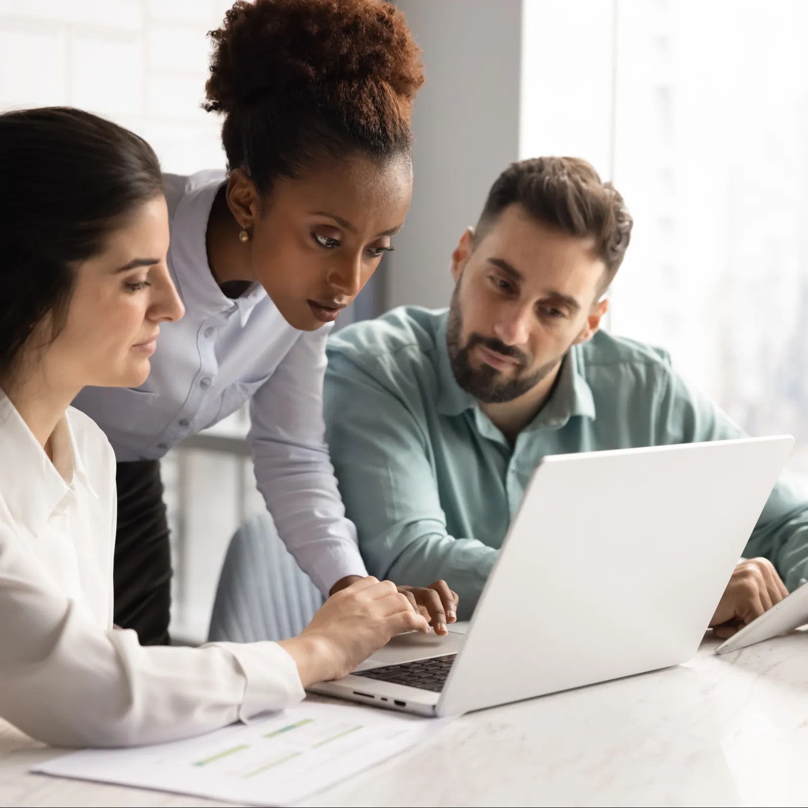 Three diverse professionals collaborating while looking at a laptop and tablet in a bright office setting