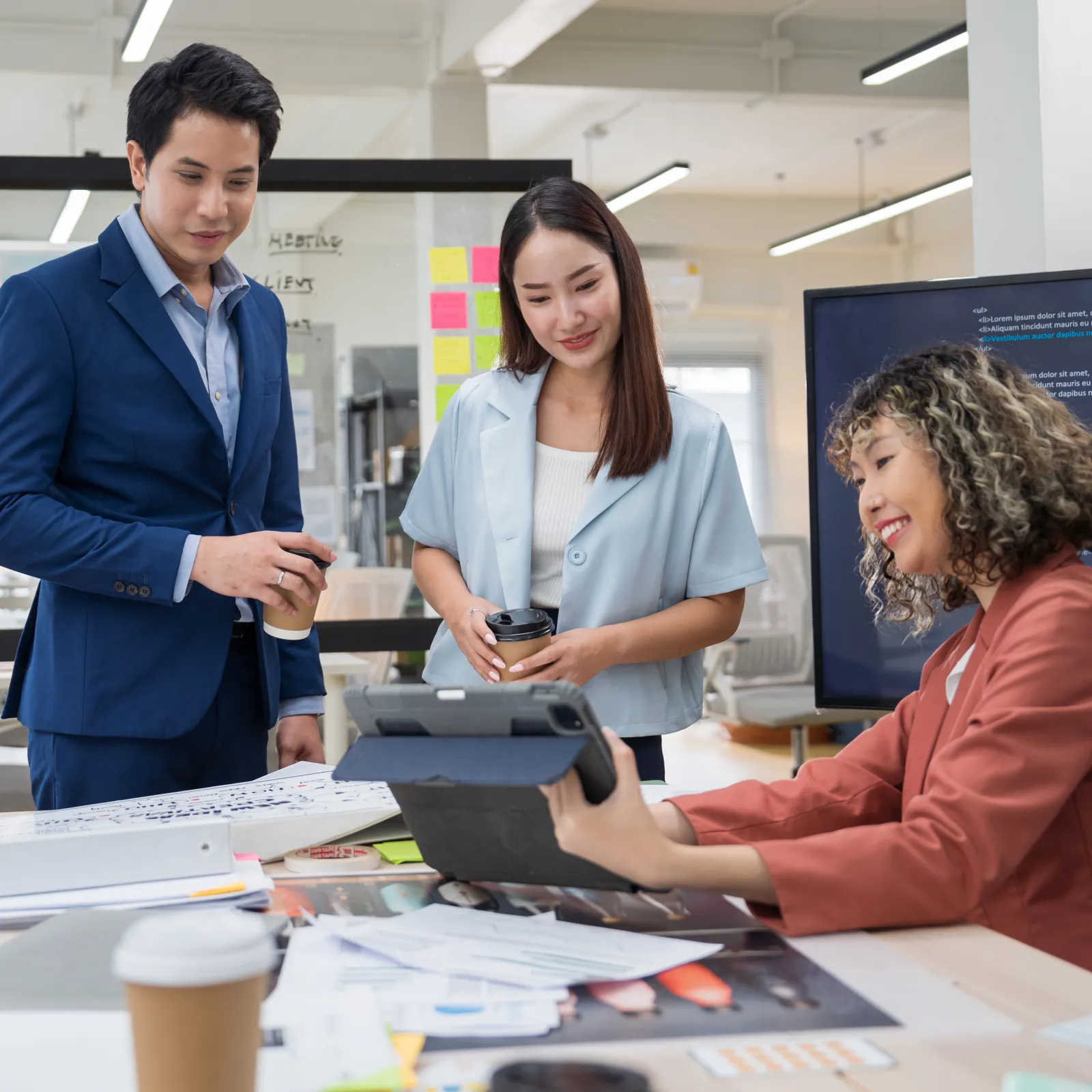 Three professionals collaborating in an office with documents, a tablet, and a screen displaying code.