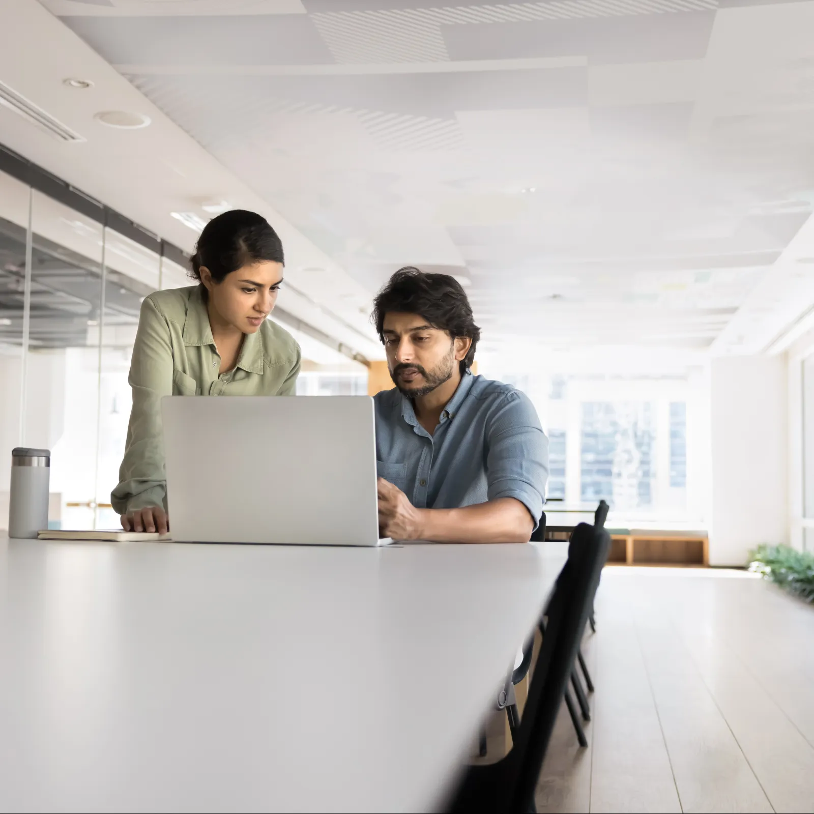Two professionals collaborating over a laptop in a modern office with large windows and glass walls.