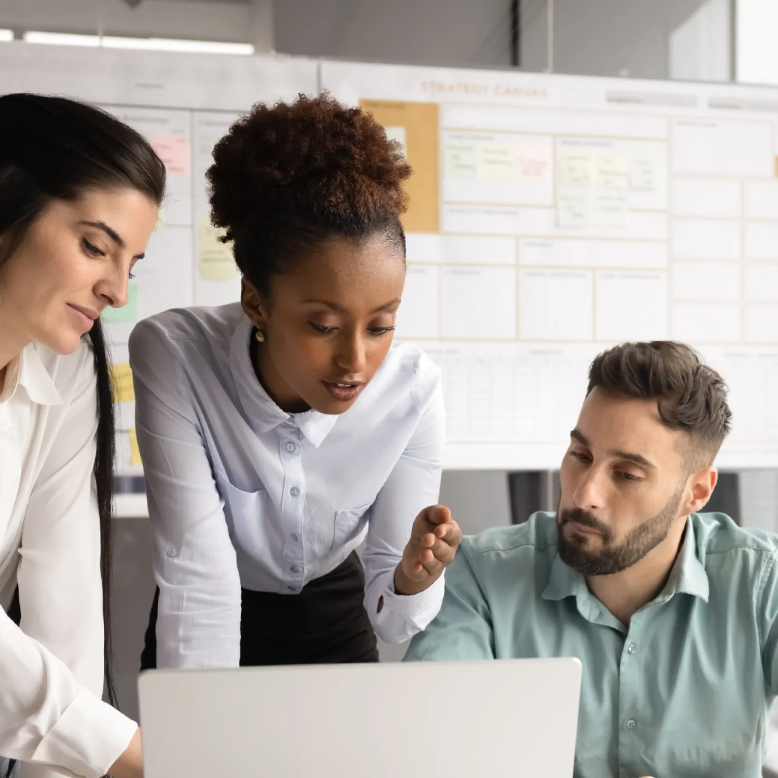 Three diverse professionals collaborating over a laptop in a modern office with business charts on glass walls.