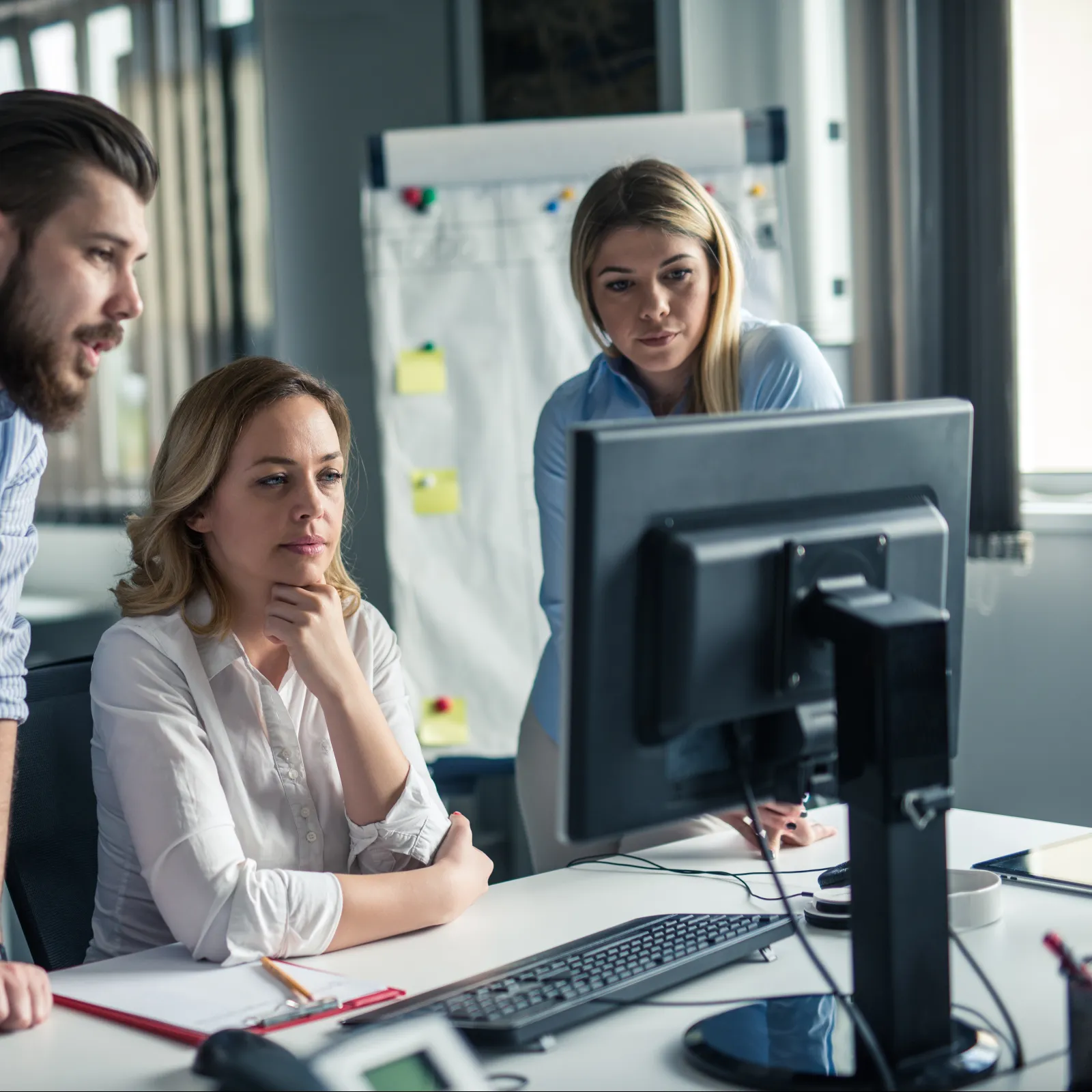 Three coworkers focused and collaborating while looking at a desktop monitor in a modern office setting.