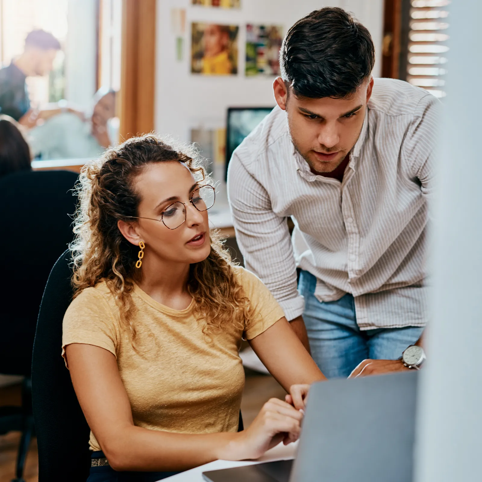 Two young professionals collaborate on a project using a laptop in a busy modern office environment.