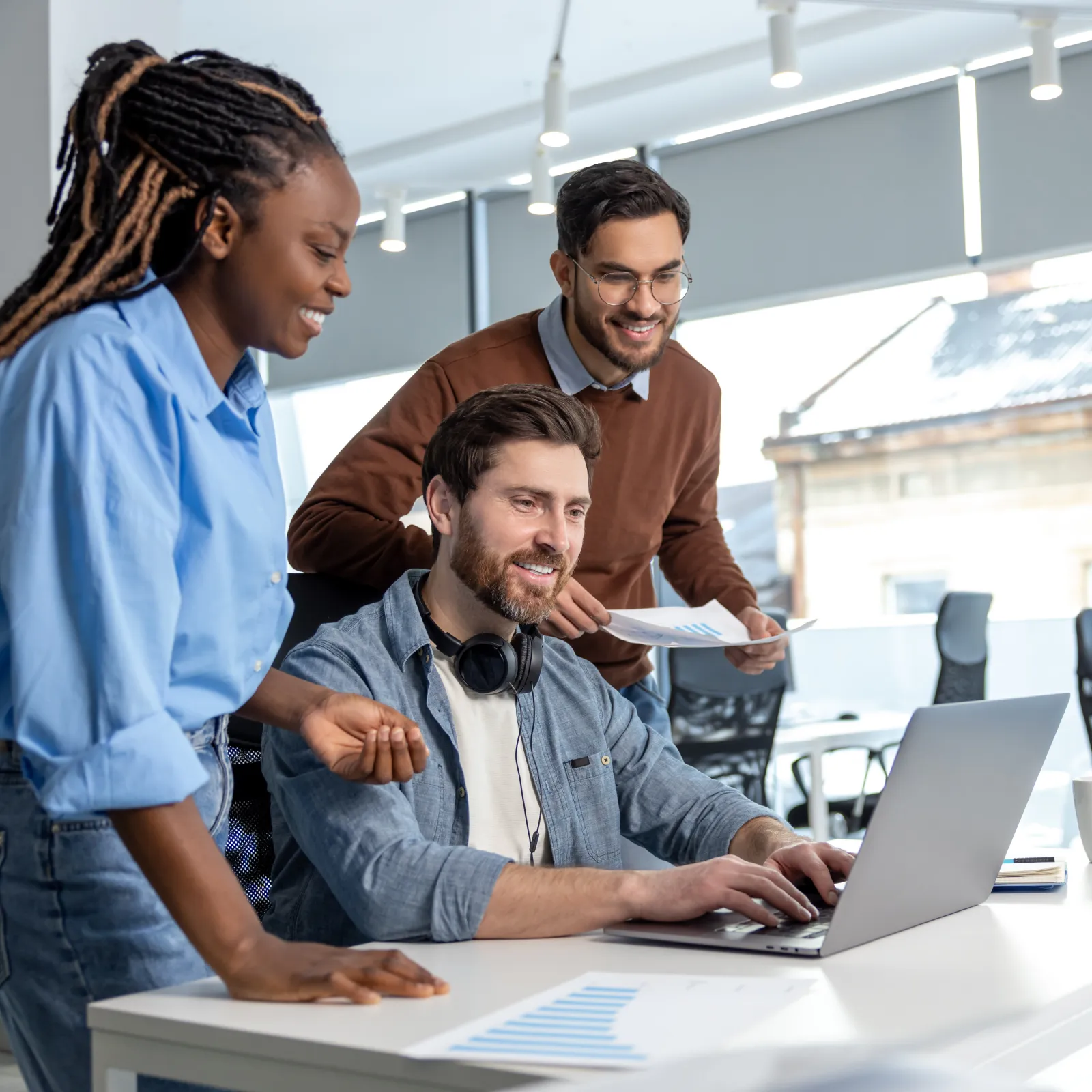 Three diverse coworkers collaborate happily around a laptop in a modern office with large windows and bright lighting.