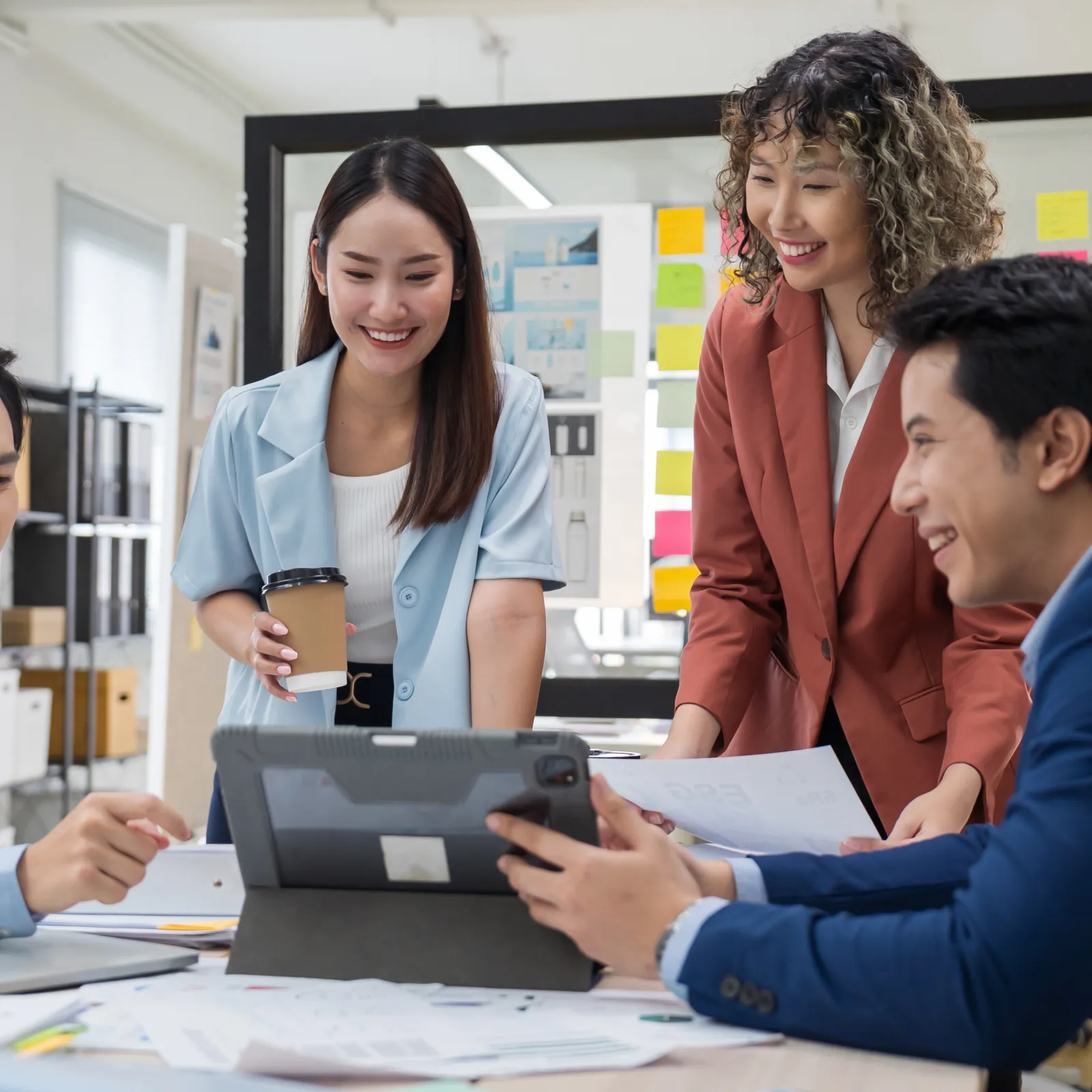 Four young professionals collaborating over a tablet in a modern office with charts and sticky notes on walls.