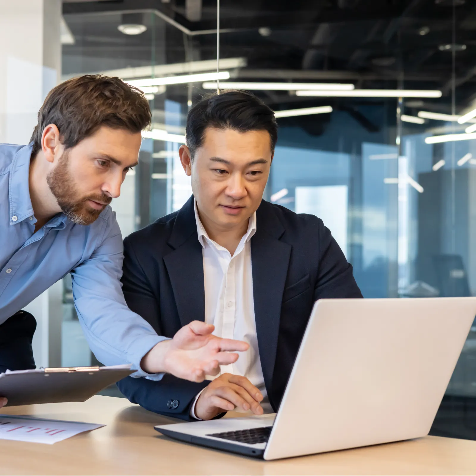 Two businessmen collaborating over a laptop in a modern office with documents and clipboard on the table