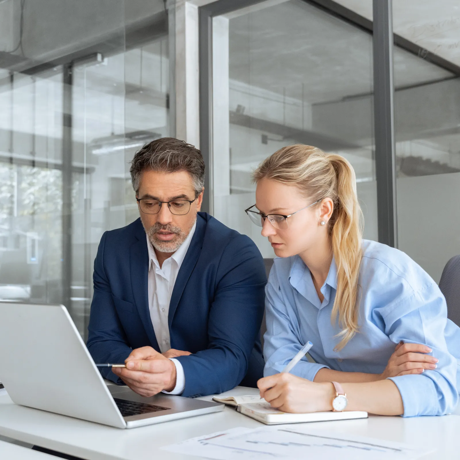 Man and woman collaborating on laptop in modern office setting, taking notes during discussion.