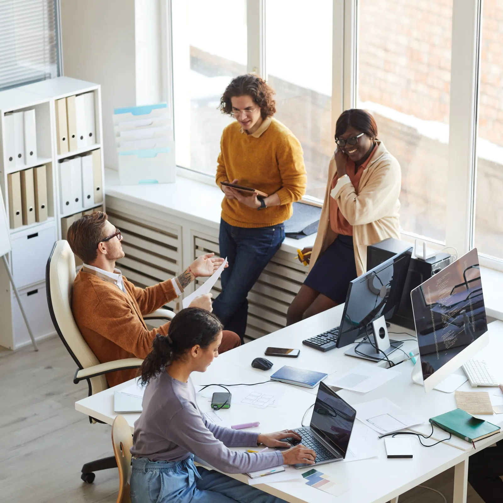 Diverse team collaborating in bright office with laptops, tablets, and large windows providing natural light.