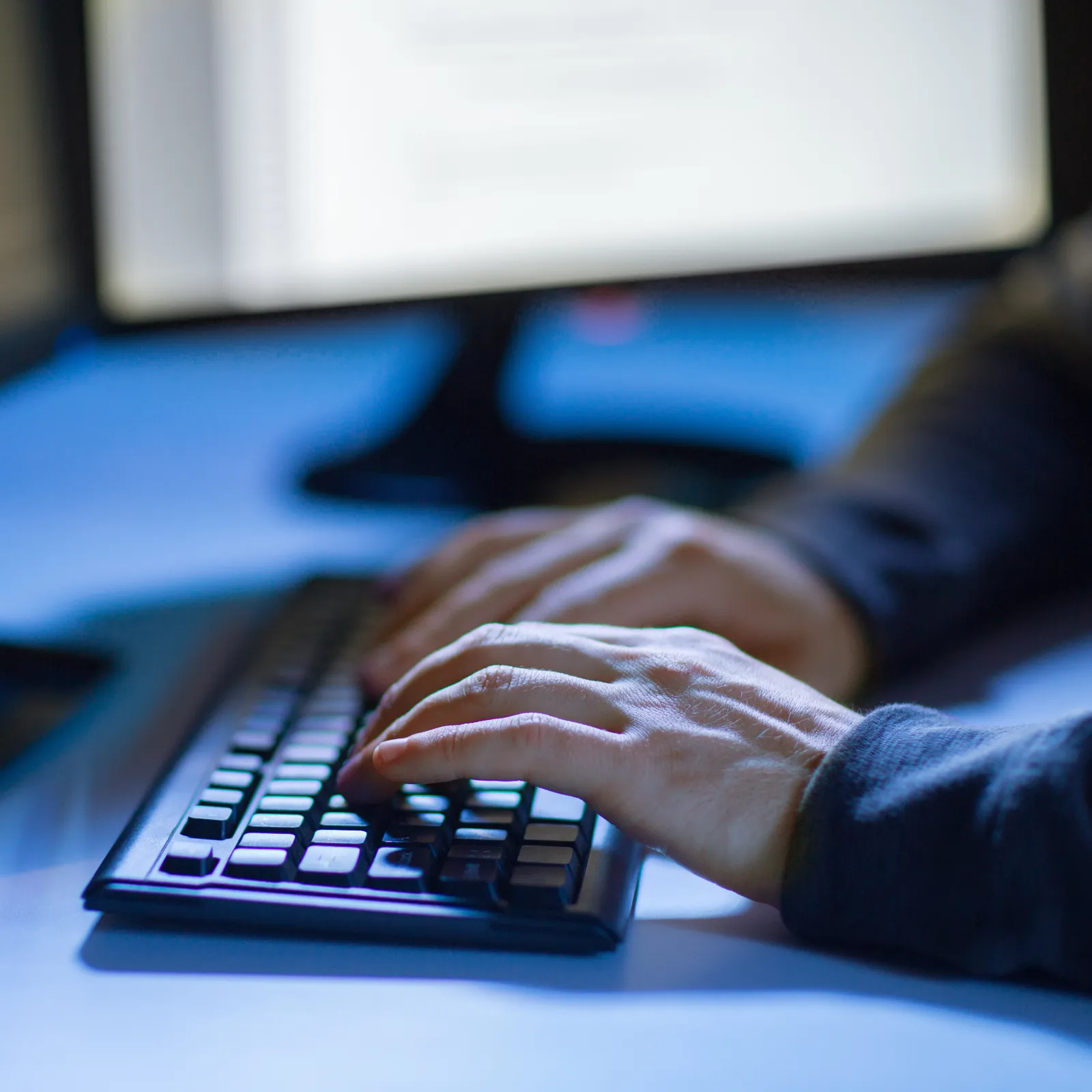 Person typing code on keyboard with dual monitors showing programming environment in dim light.