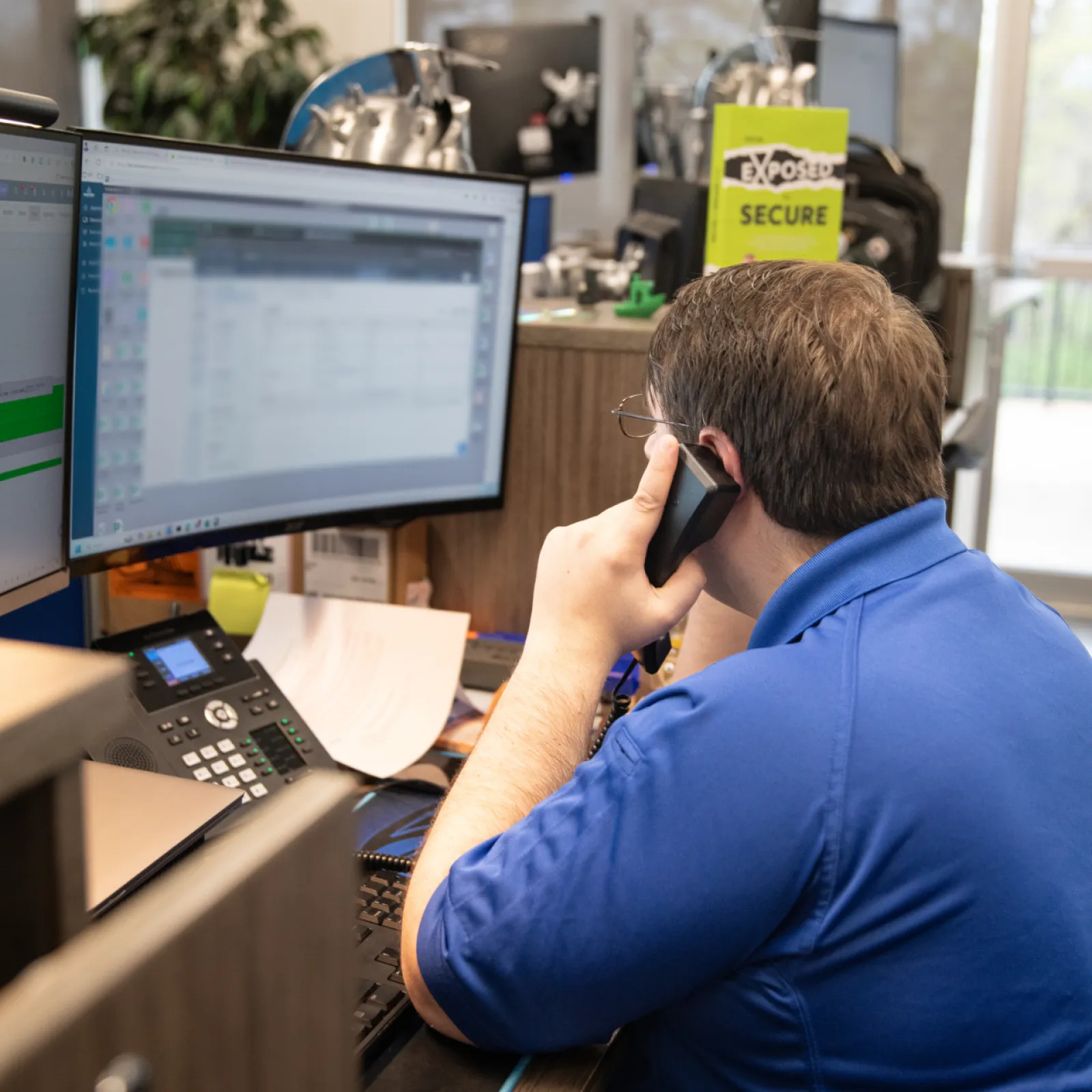 An employee in a blue shirt working at a desk with computers and a phone, focused on a task.