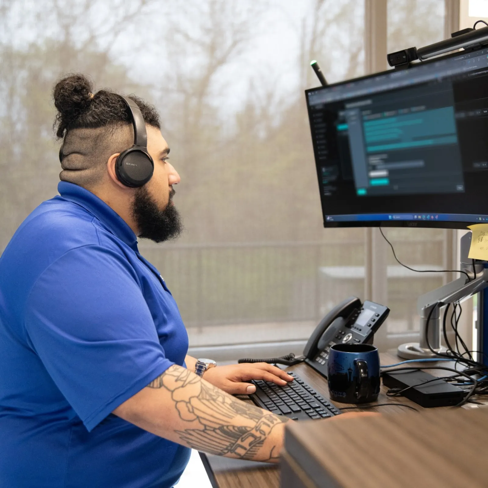 Man in blue shirt using a computer with headphones in a modern office setting.