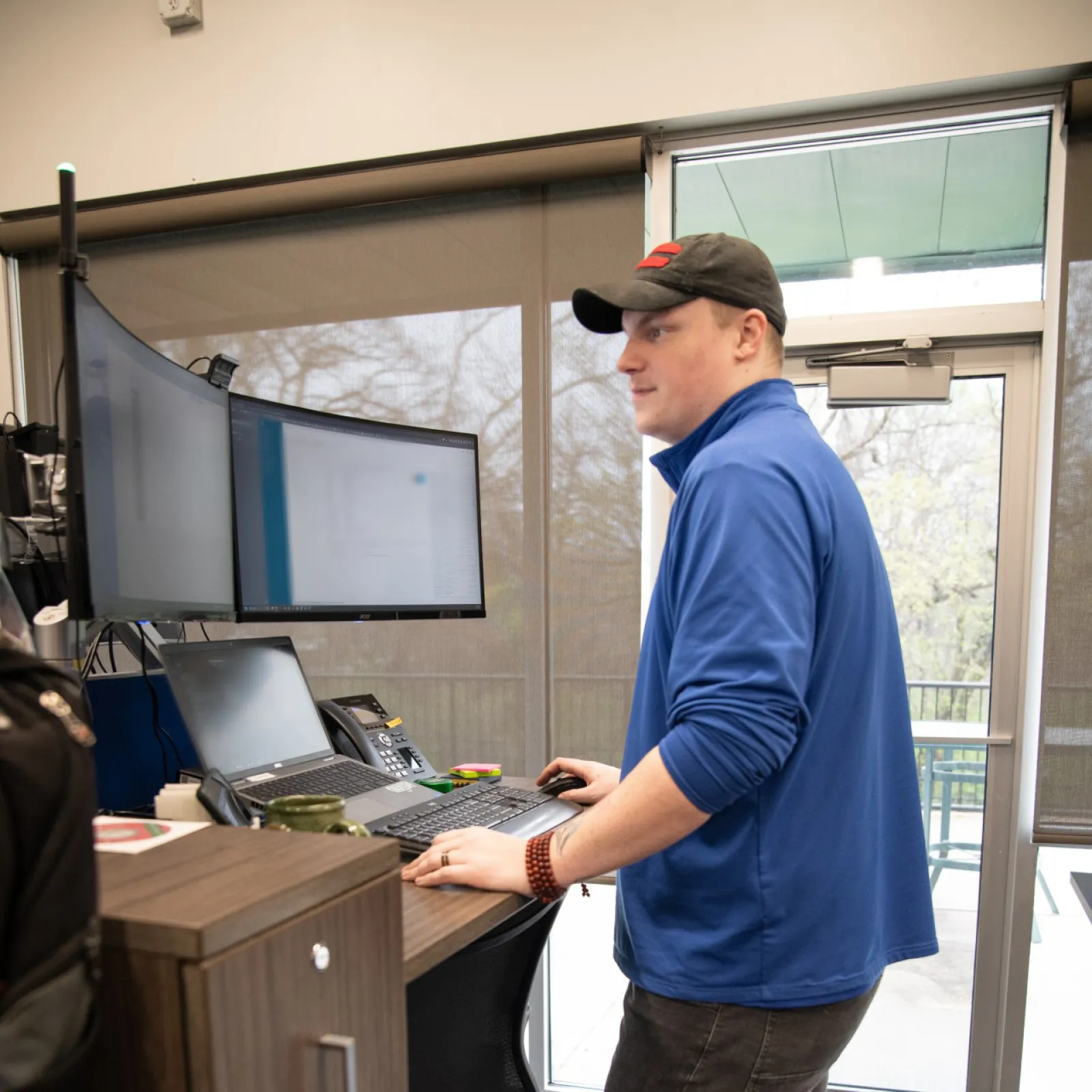 Man in blue jacket and black cap working on dual monitors at standing desk in modern office space