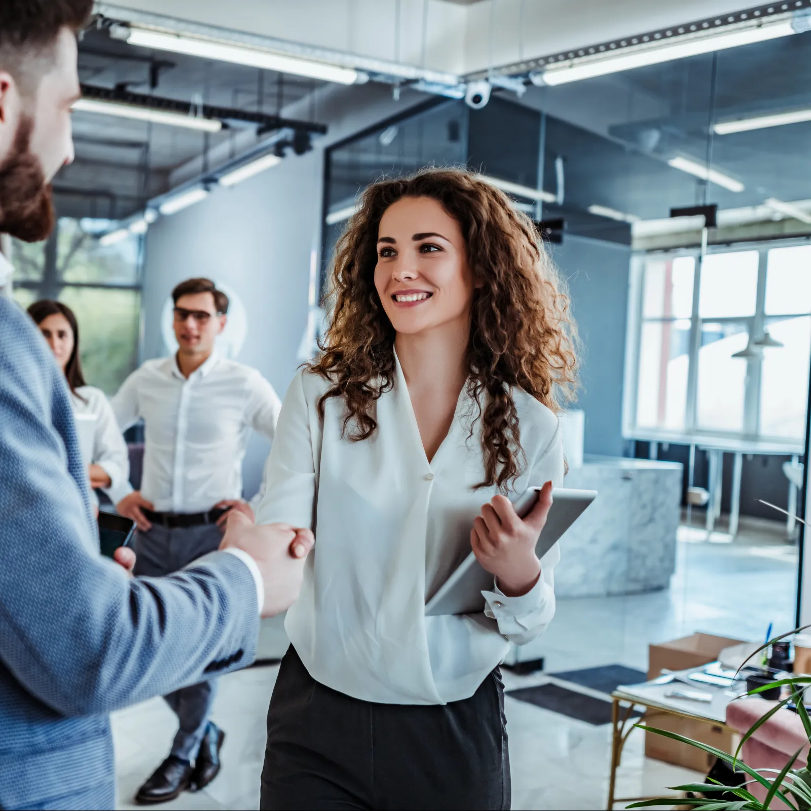 Business professionals shaking hands in modern office with colleagues in background during a meeting.