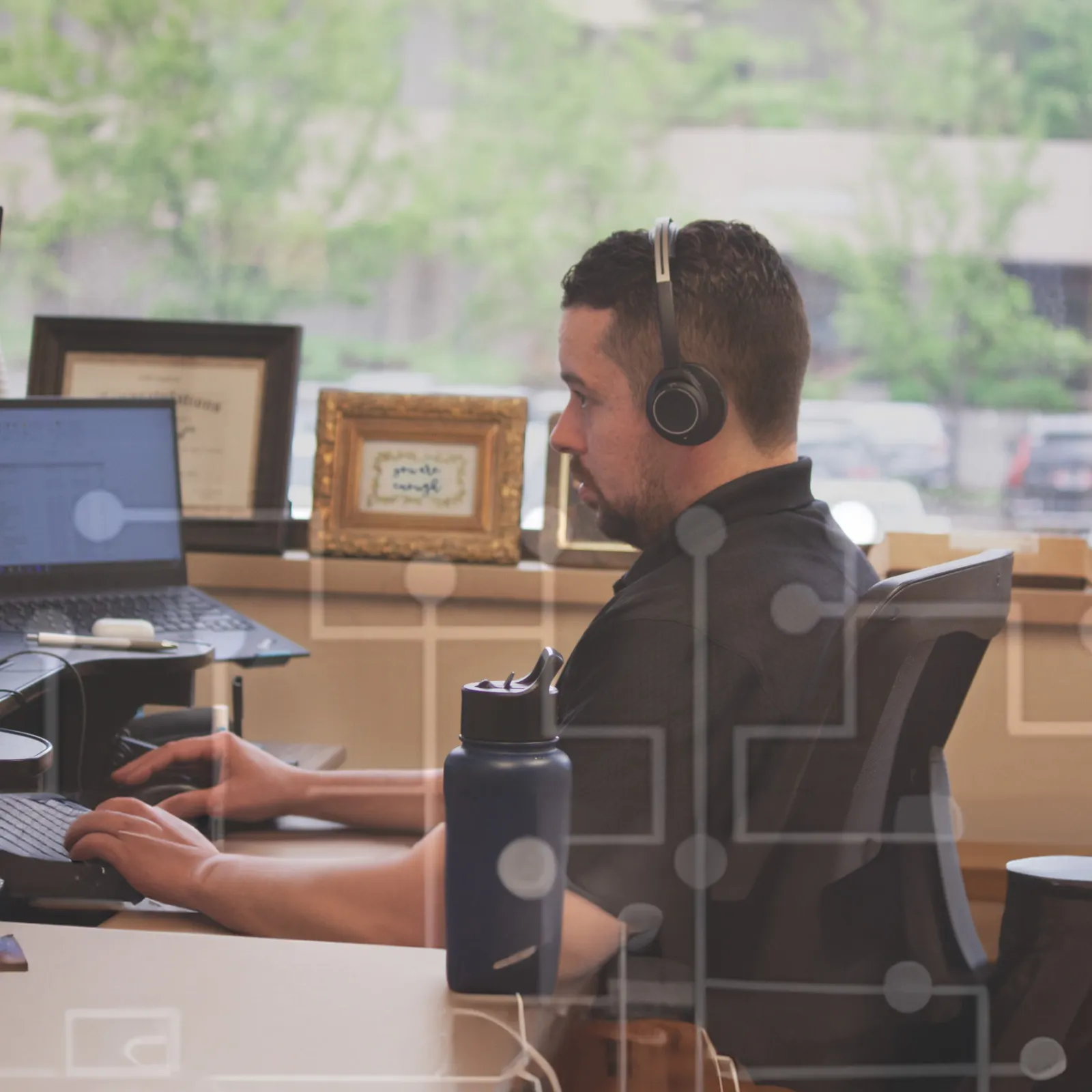 Man wearing headphones working on laptop at desk with framed photos and water bottle in bright office.