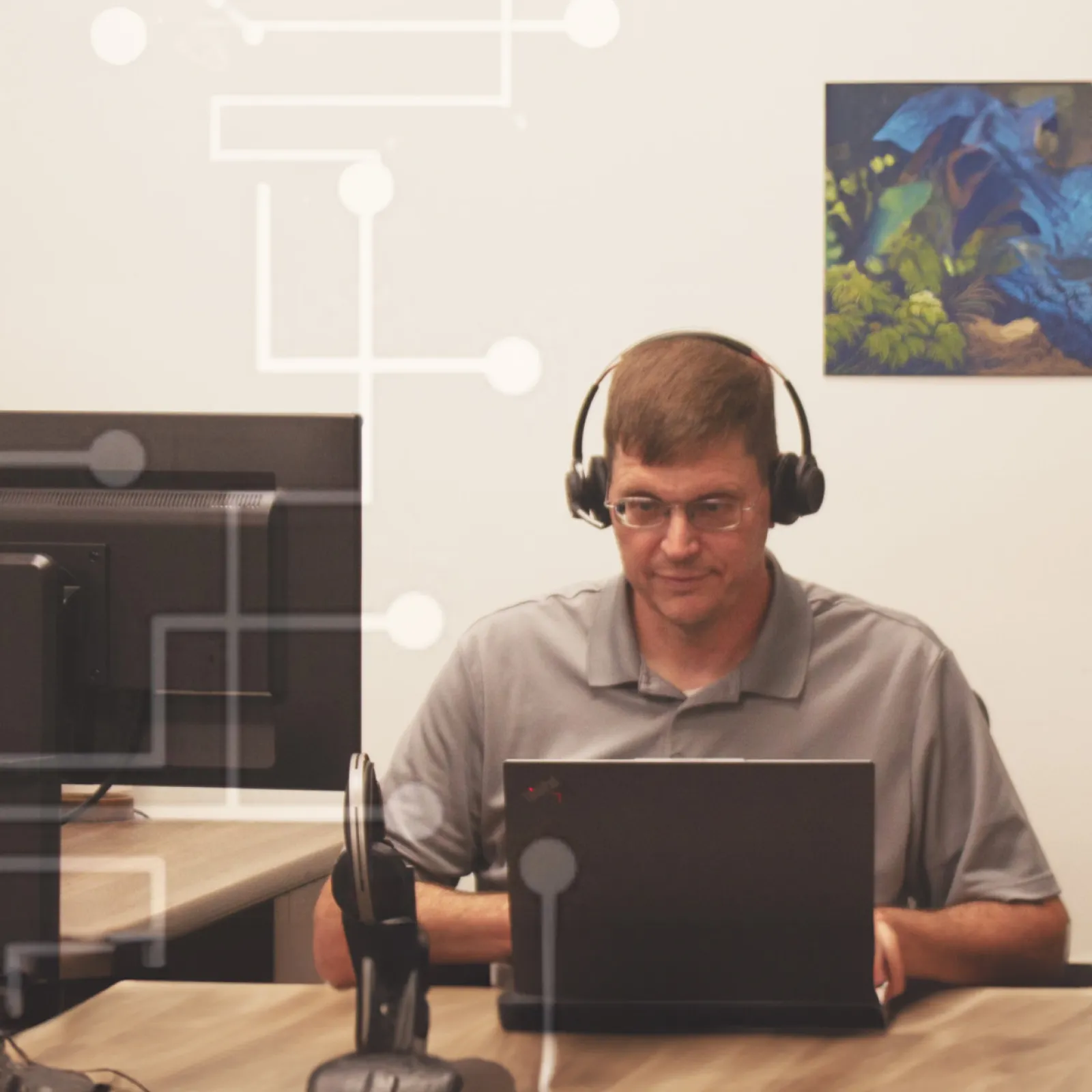 Man wearing headphones working on a laptop at a wooden desk in an office with monitors and abstract tech graphics.