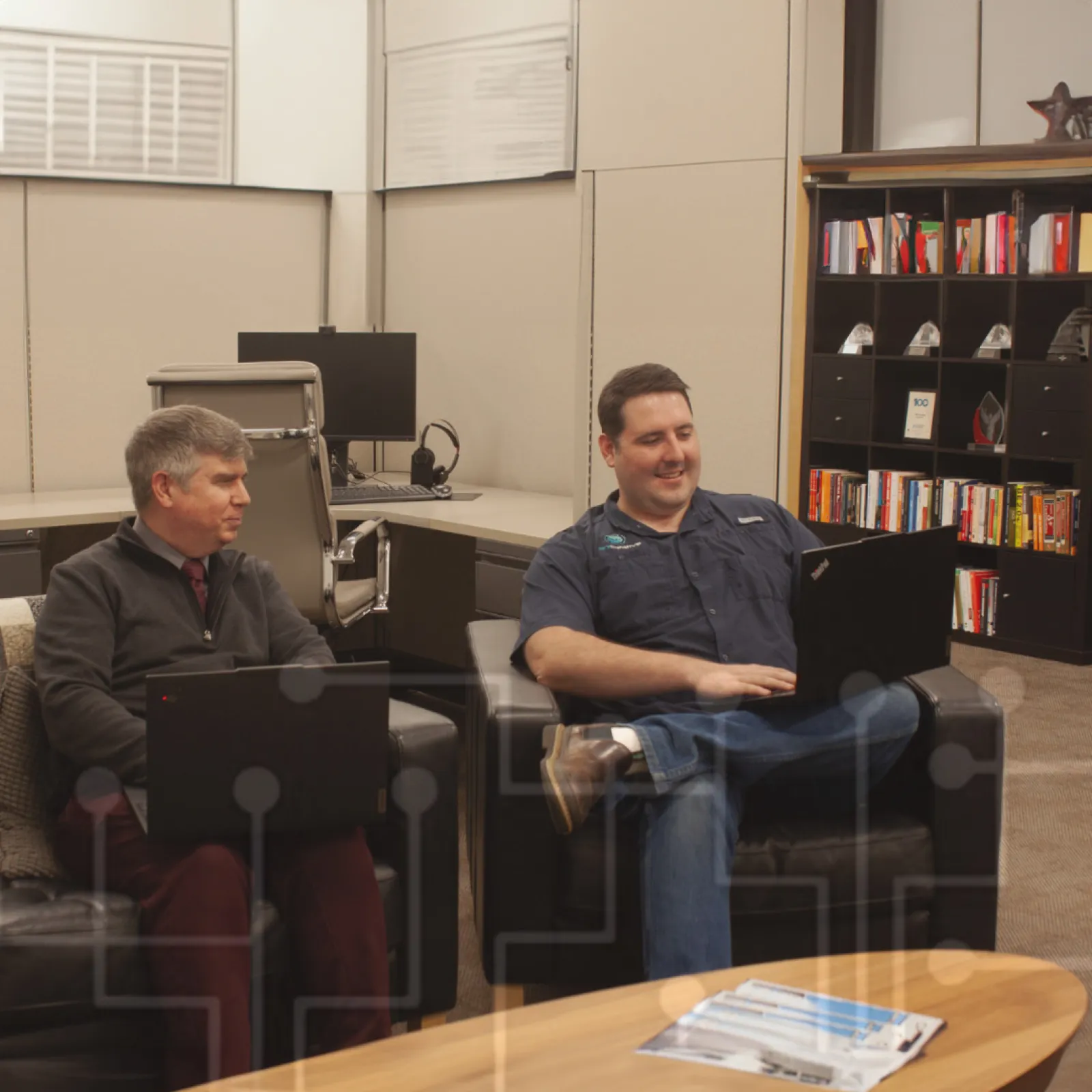 Two men sitting on black chairs in an office lounge working and chatting with laptops open.