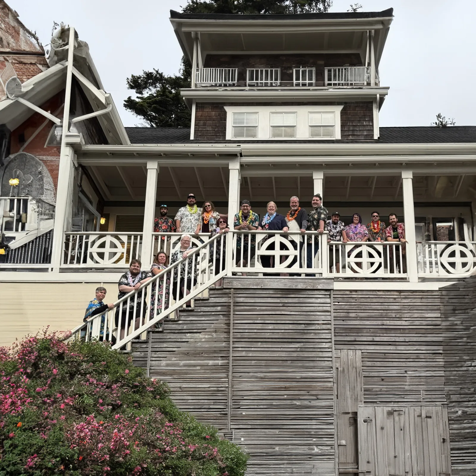 Group of people wearing colorful leis standing on the porch of a large wooden house with flowers below.