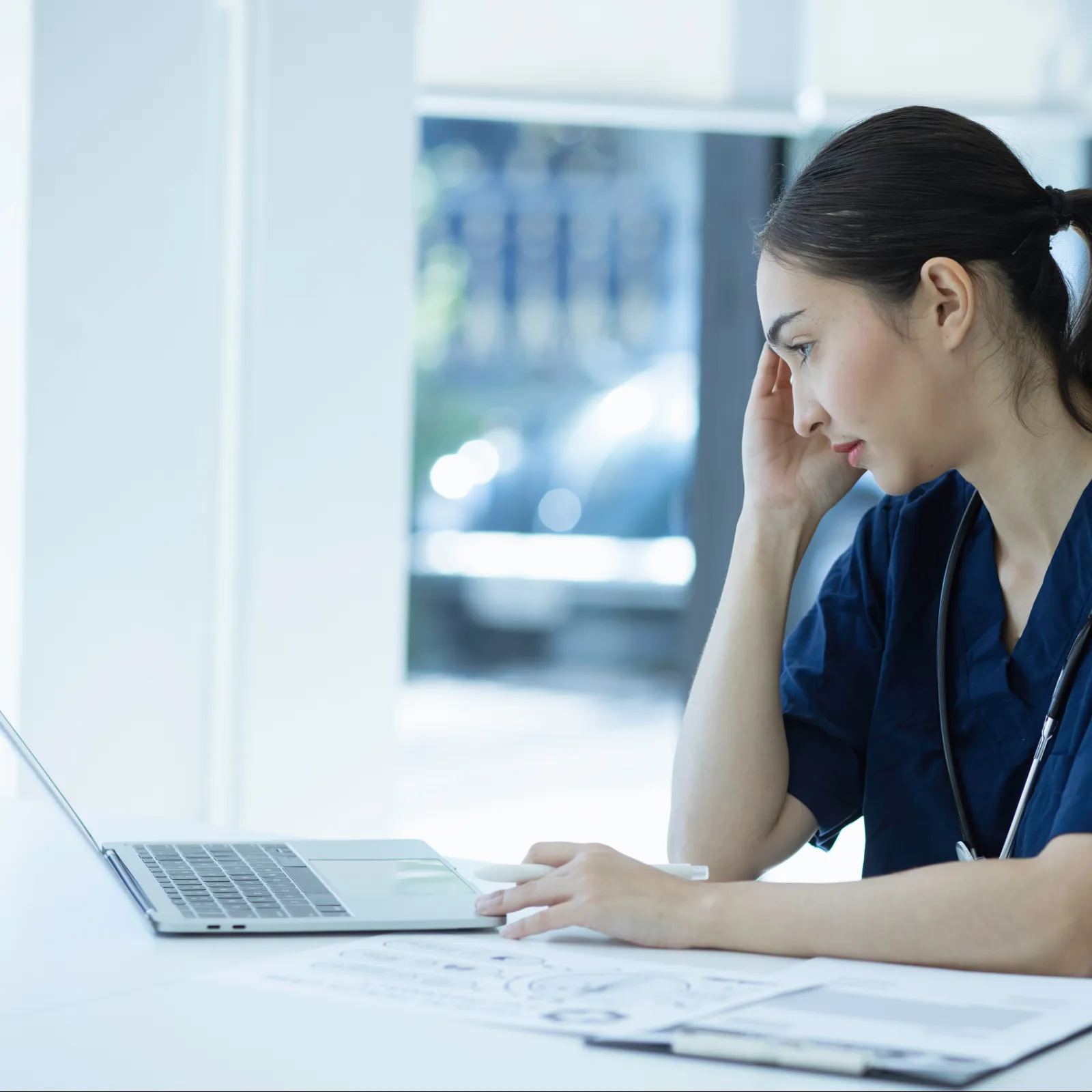 Female healthcare professional with stethoscope using laptop and reviewing documents in bright office workspace.