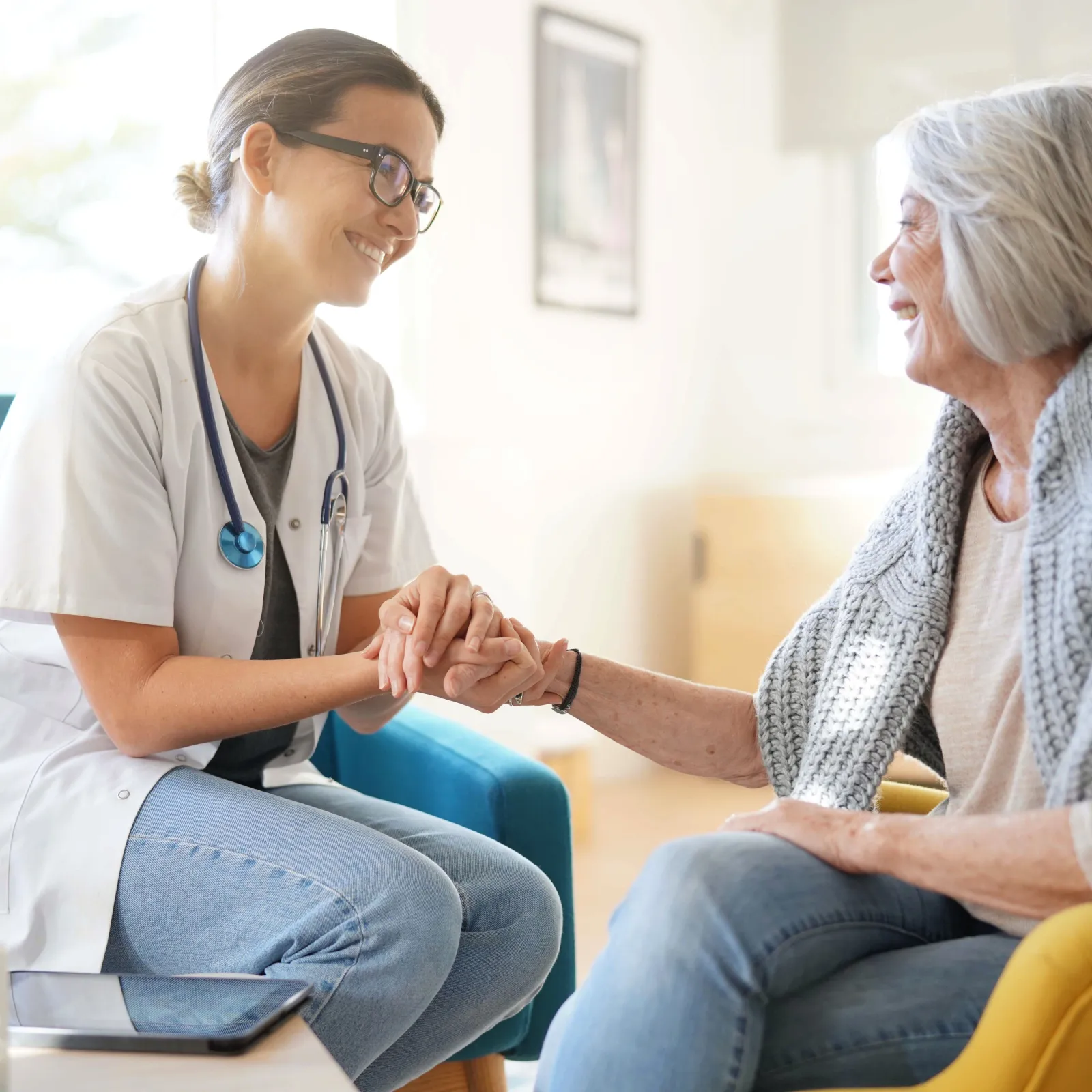 Young female doctor holding hands and smiling at elderly woman in cozy room during consultation.