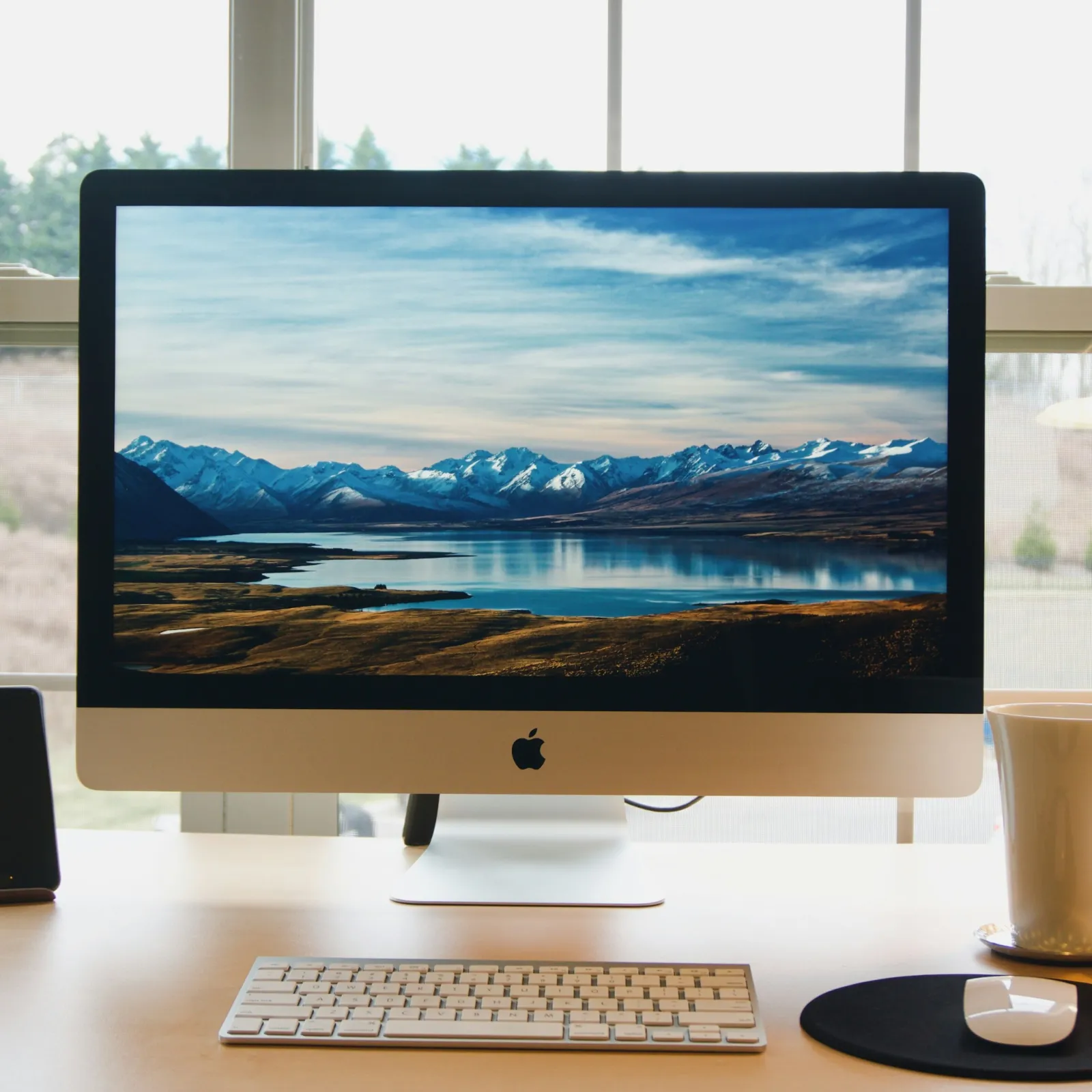 A sleek Apple computer displaying a scenic mountain lake view beside a workspace setup.