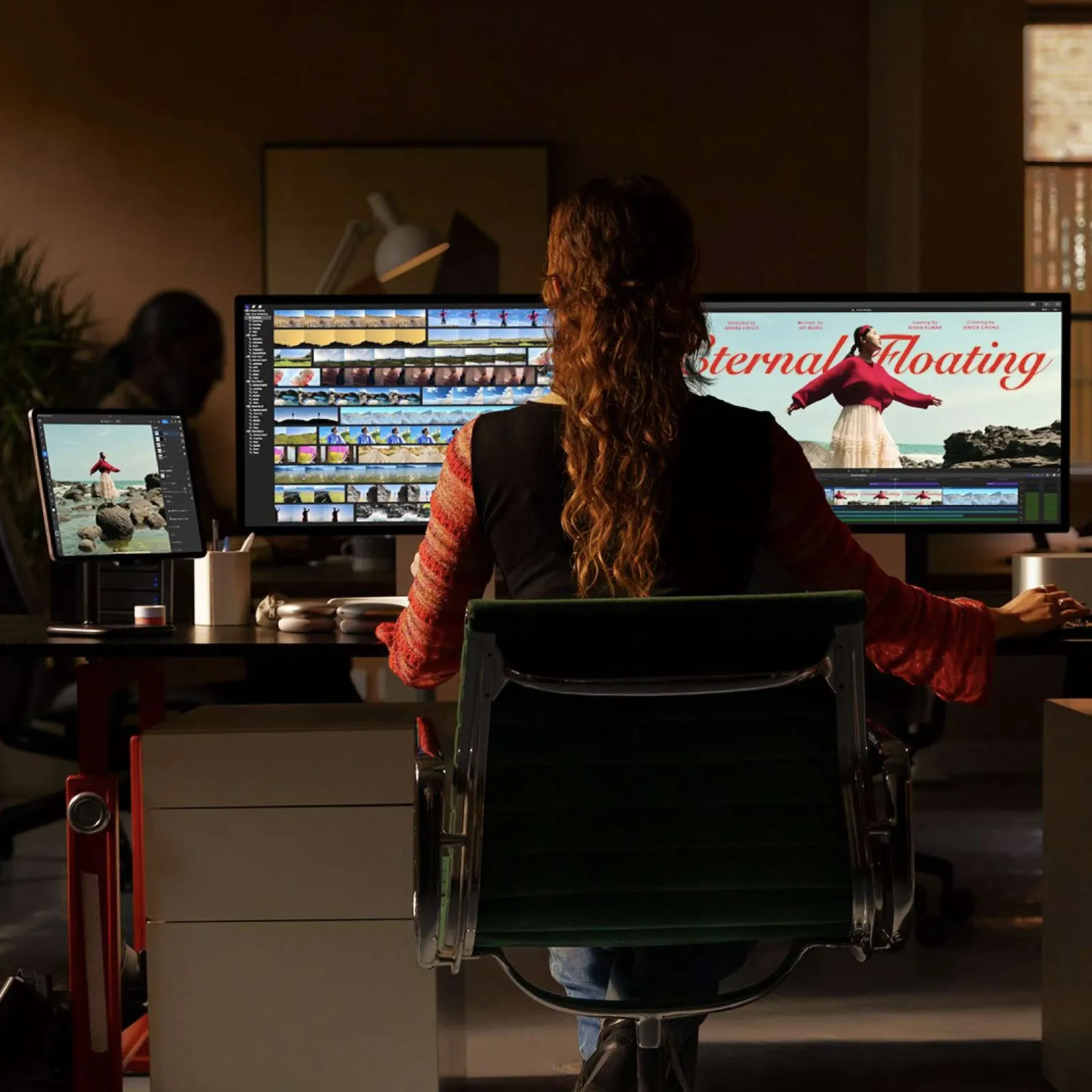 Person editing video on a large dual-monitor setup in a dimly lit modern office workspace.