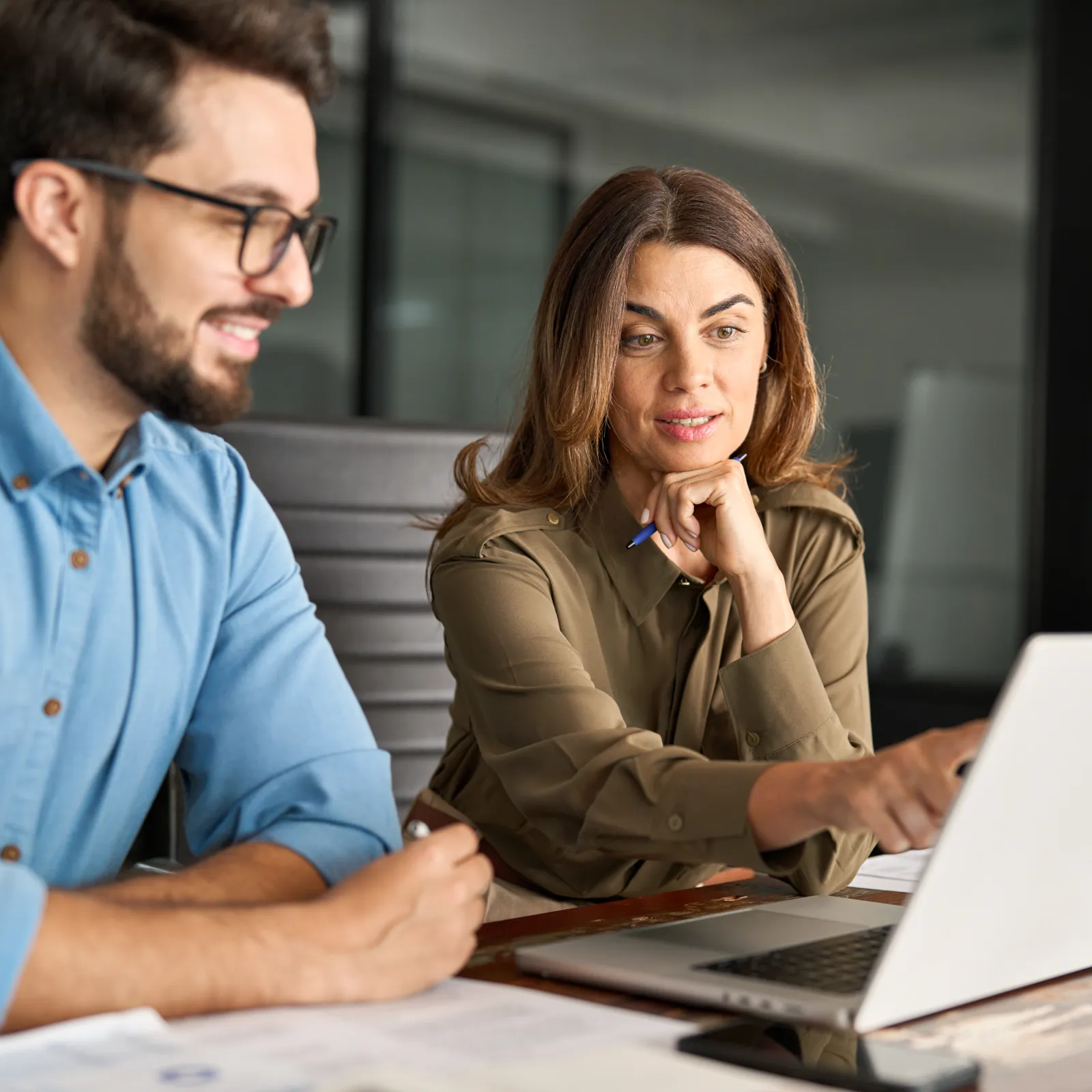 Two coworkers collaborating and discussing work on a laptop in a modern office setting.