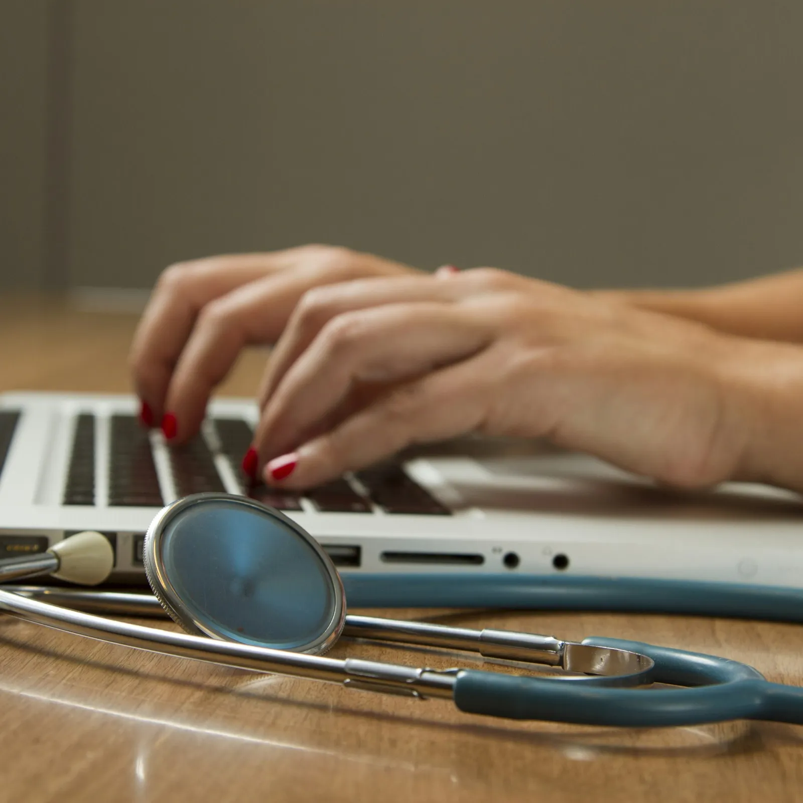 A person's hands typing on a laptop beside a stethoscope on a wooden table.