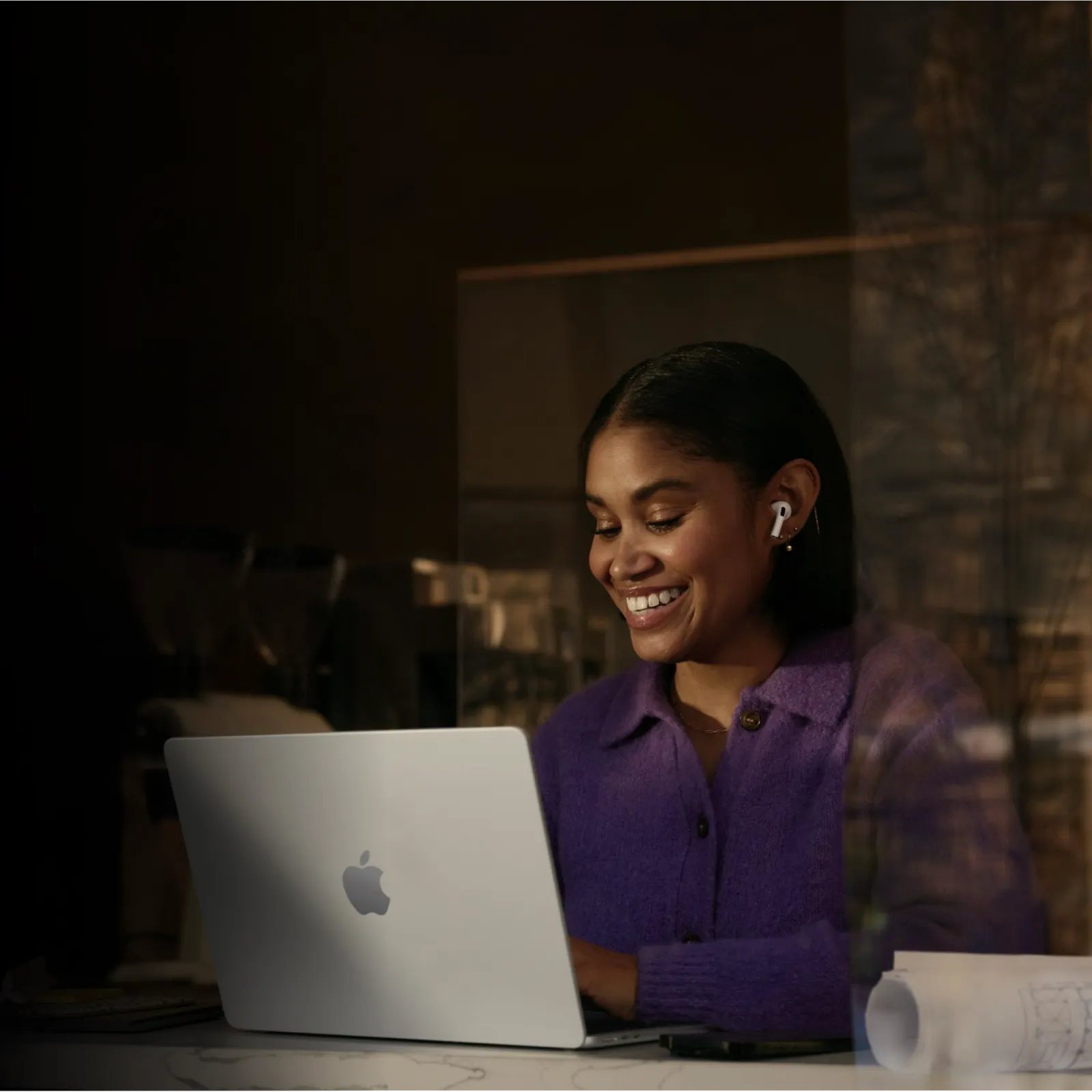 Smiling woman in purple sweater working on a MacBook with wireless earbuds in a cozy indoor setting.