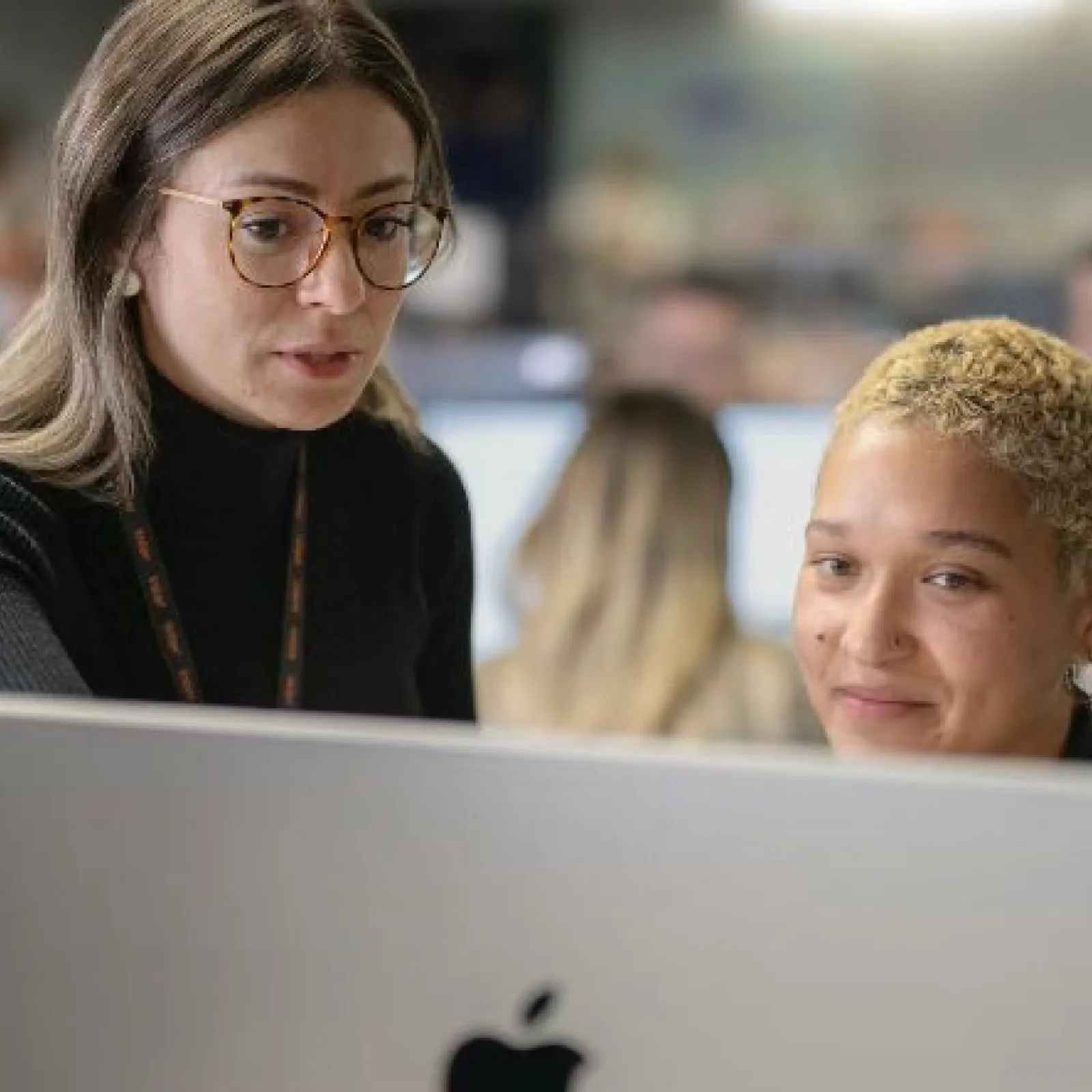Two colleagues collaborate and discuss work on a large Apple desktop computer in a modern office setting.