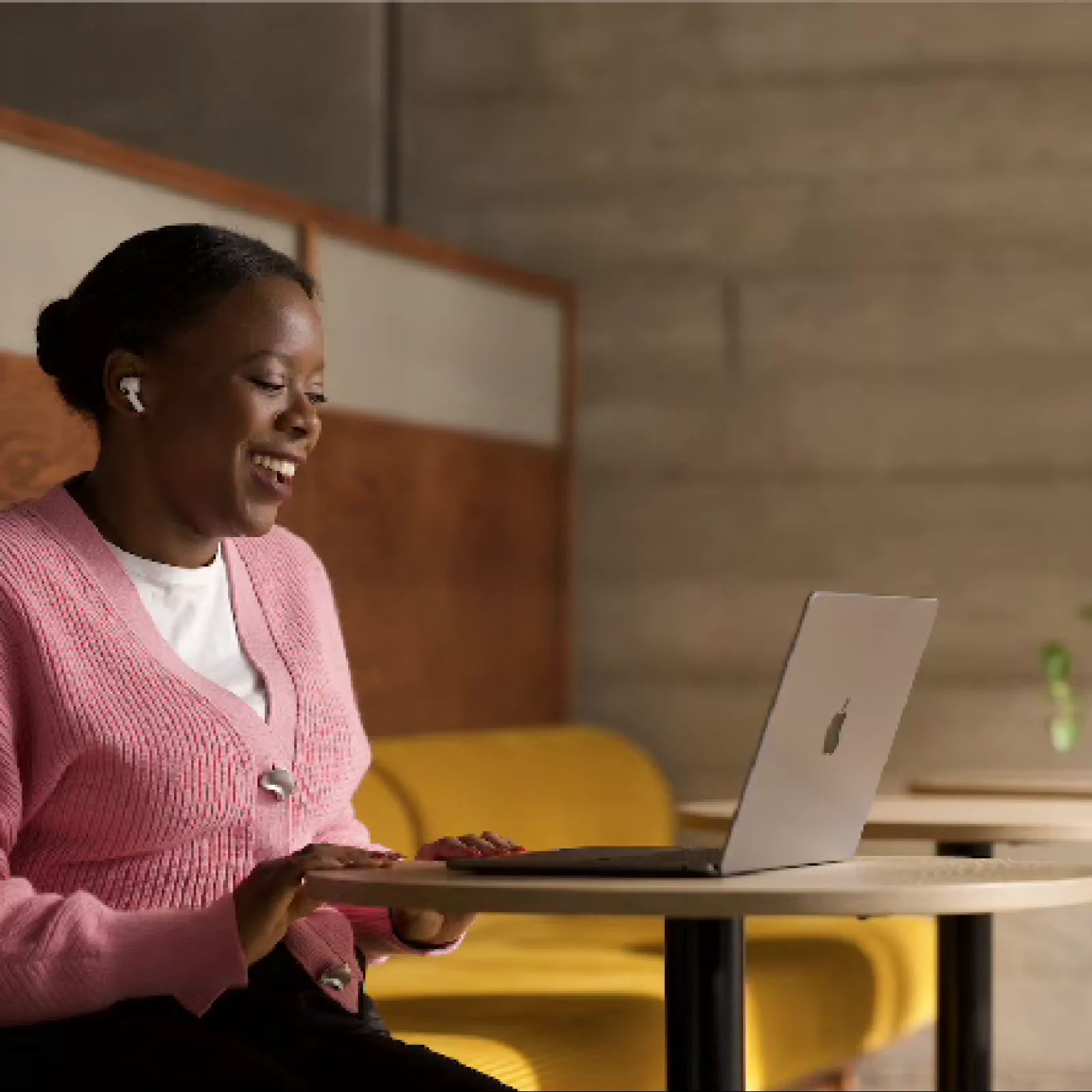 Woman in pink sweater using laptop and wireless earbuds while sitting on yellow bench in modern cafe.