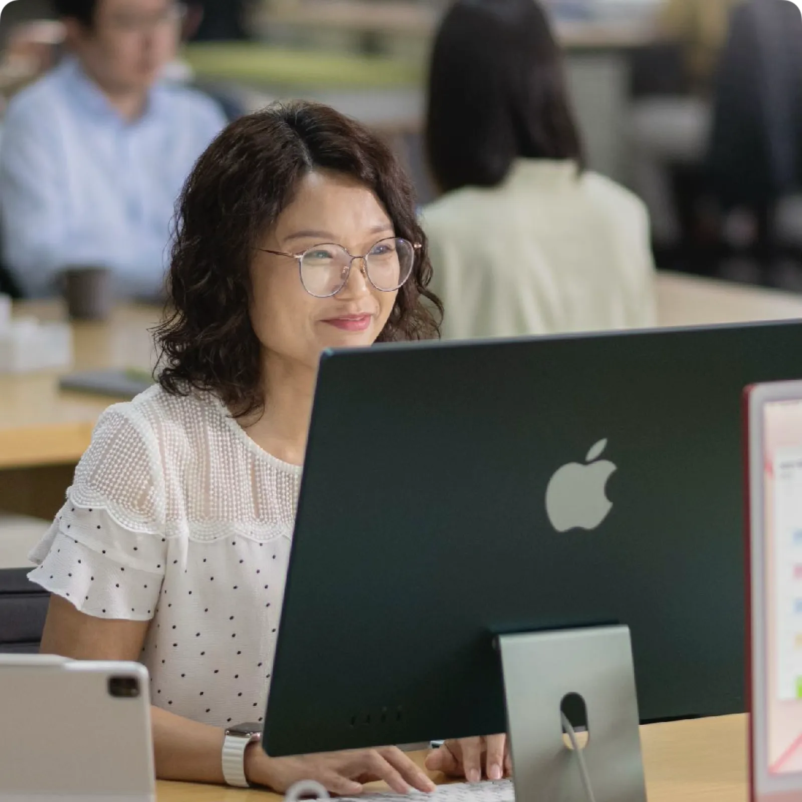 Woman wearing glasses working on an Apple iMac desktop computer in a modern office setting.