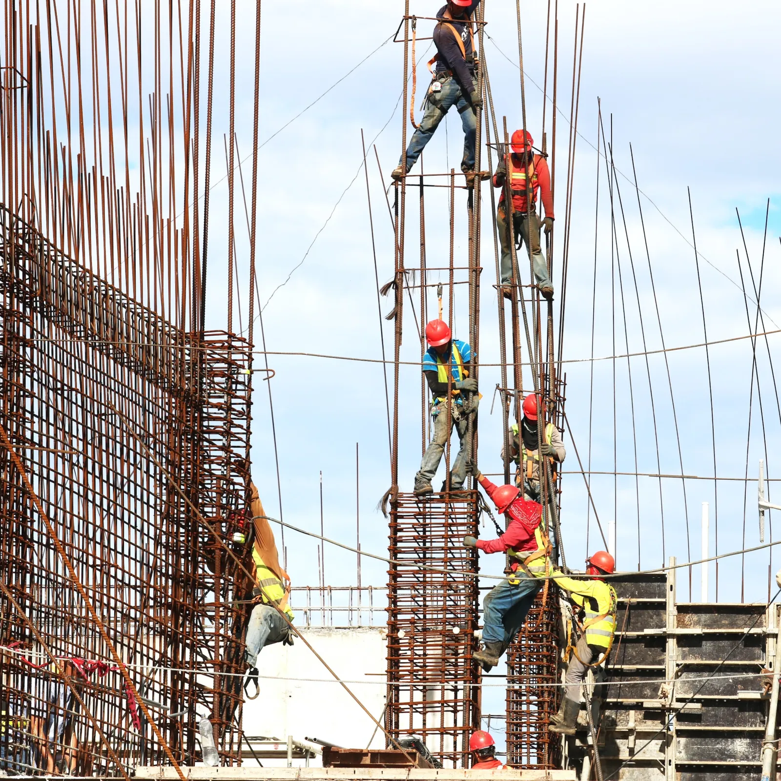 Construction workers wearing safety gear working on steel rebar framework at a building site under clear sky.