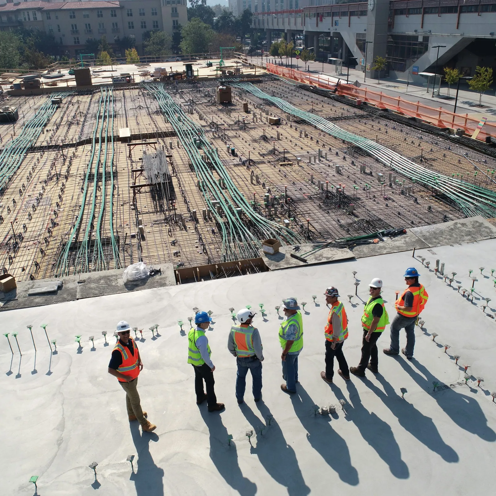 Construction workers in safety gear inspecting a large building site with exposed pipes and wiring beneath a concrete slab.