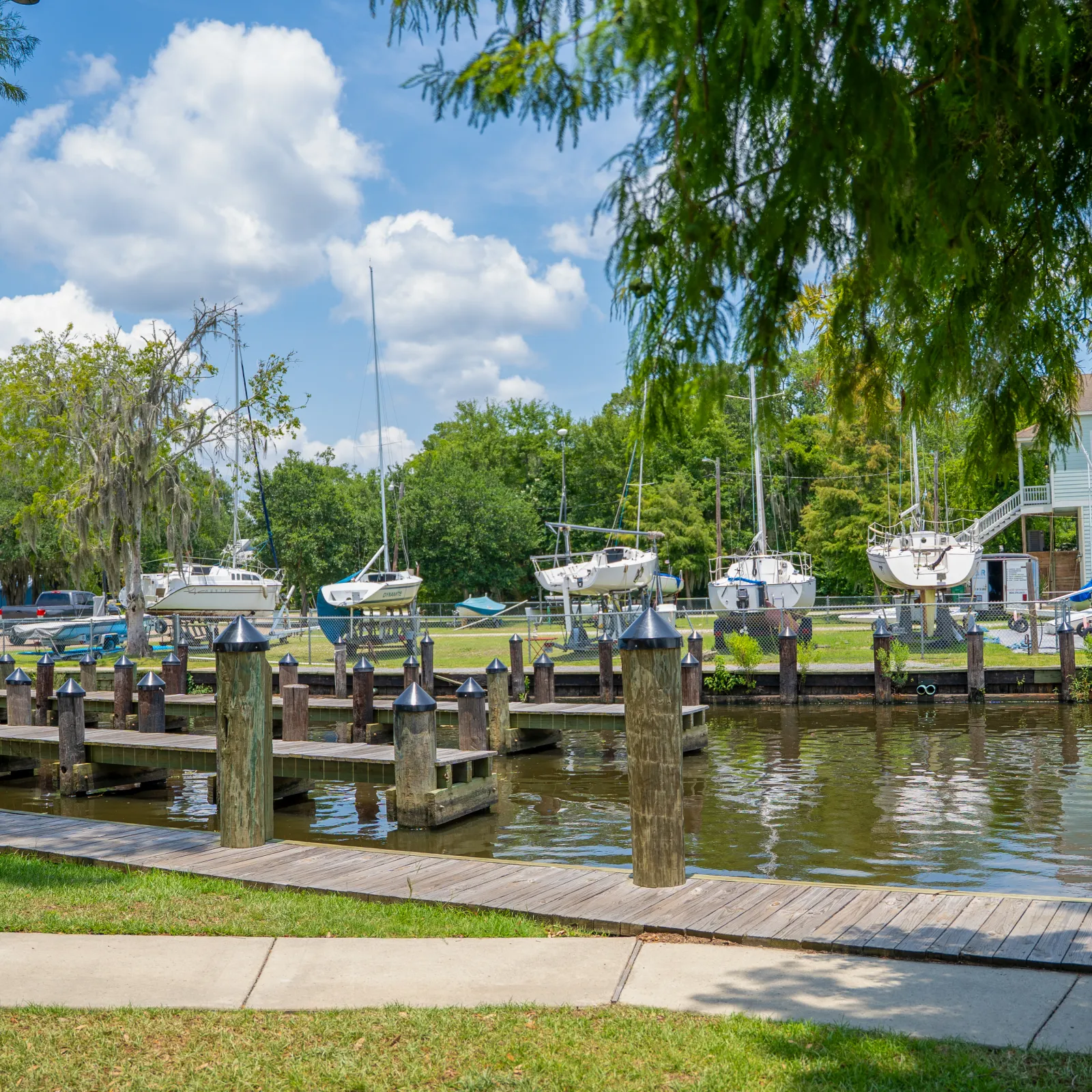Sunny marina with docked sailboats, wooden piers, green trees, and blue sky with white clouds.