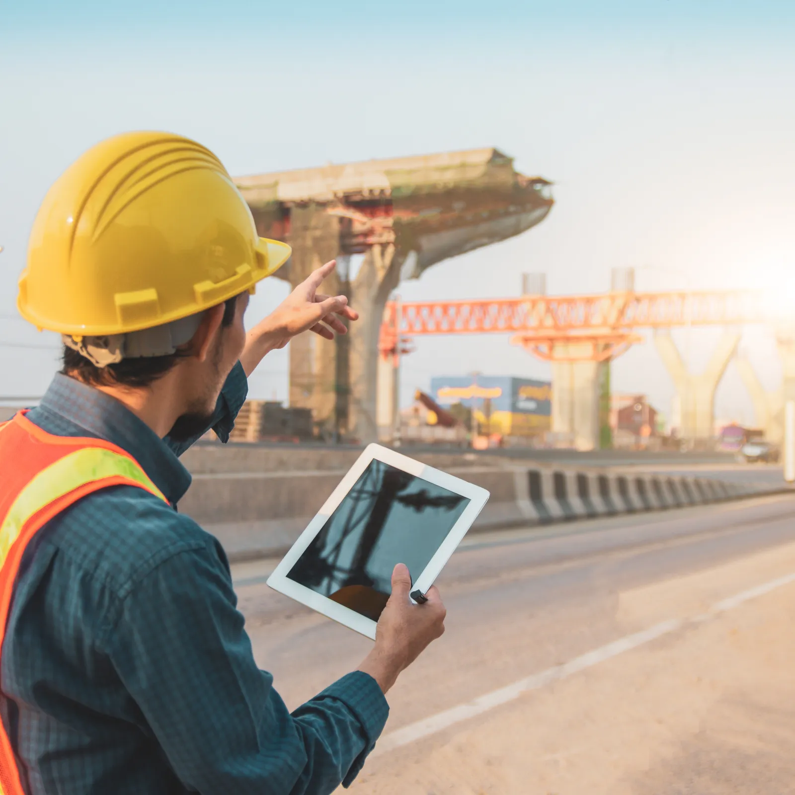 Construction worker in safety gear using tablet and pointing at bridge under construction on sunny day