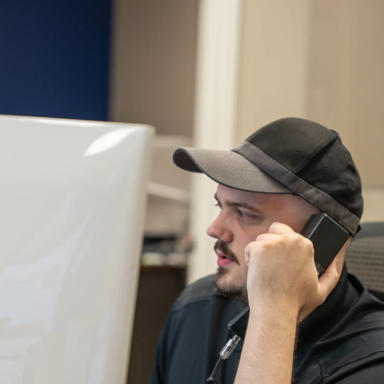 Man wearing a black cap talking on the phone while working at a computer in an office environment