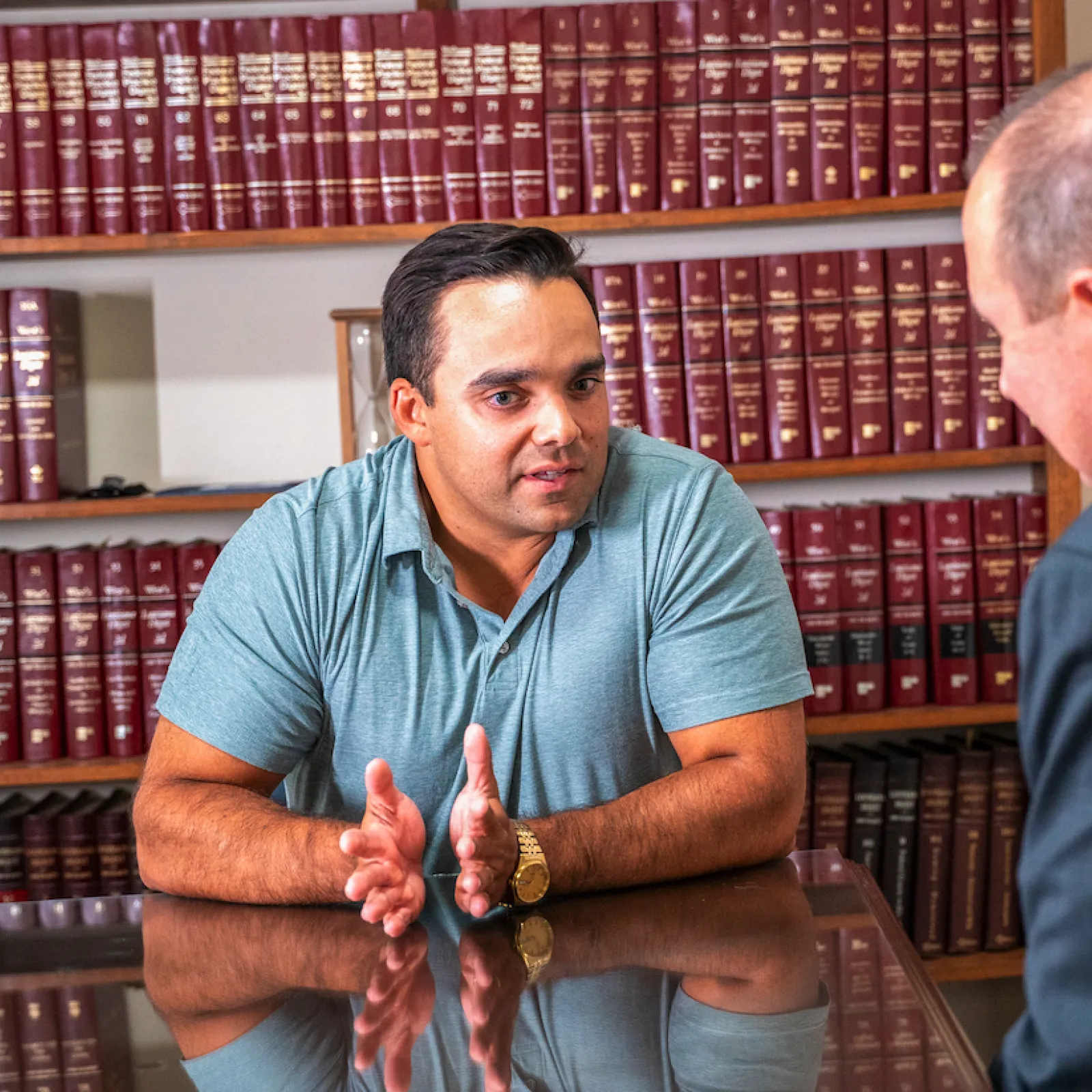 Two men engaged in discussion at a table in a room lined with red law books and shelves.