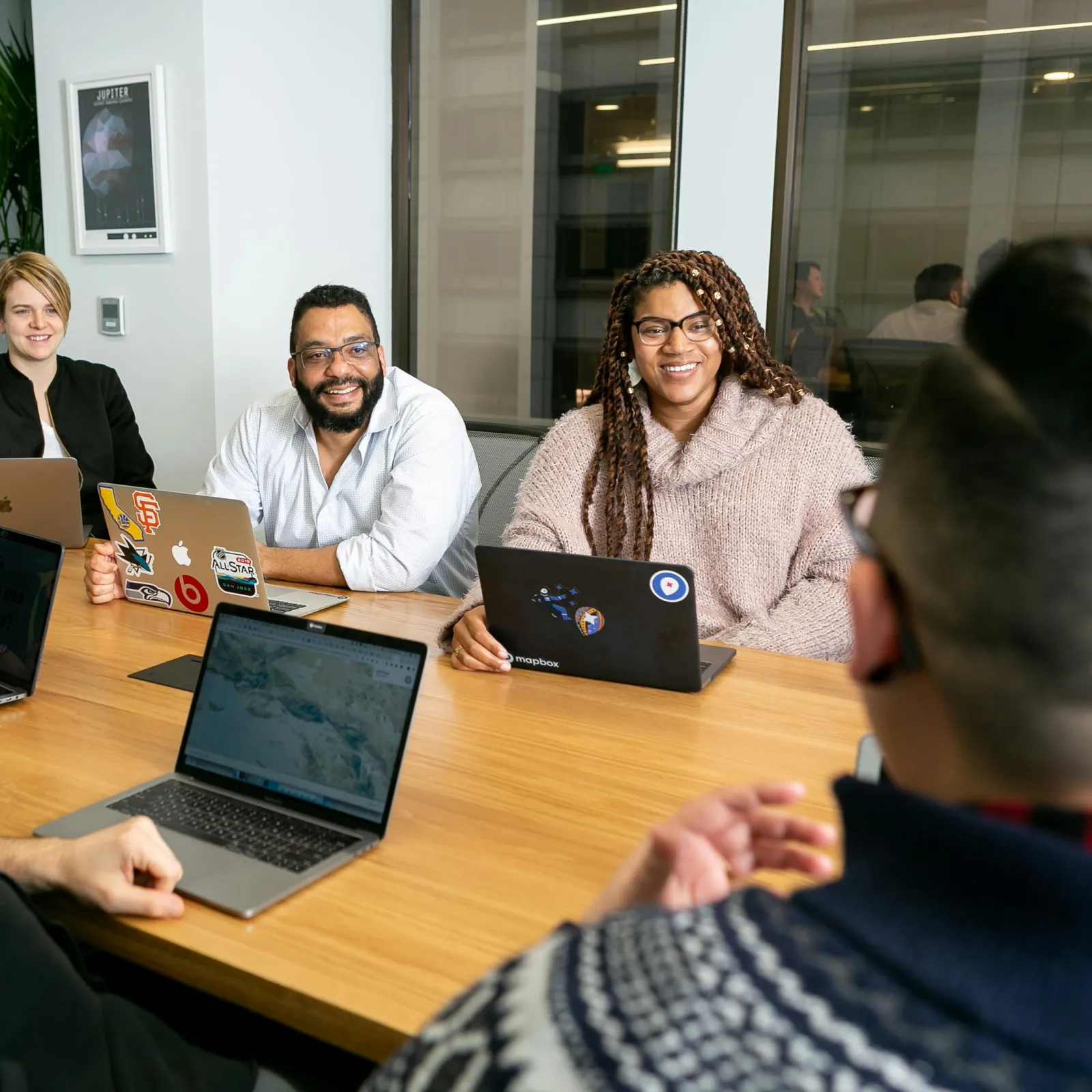 Diverse group of five coworkers engaging in a meeting around a wooden table with laptops and smiles.
